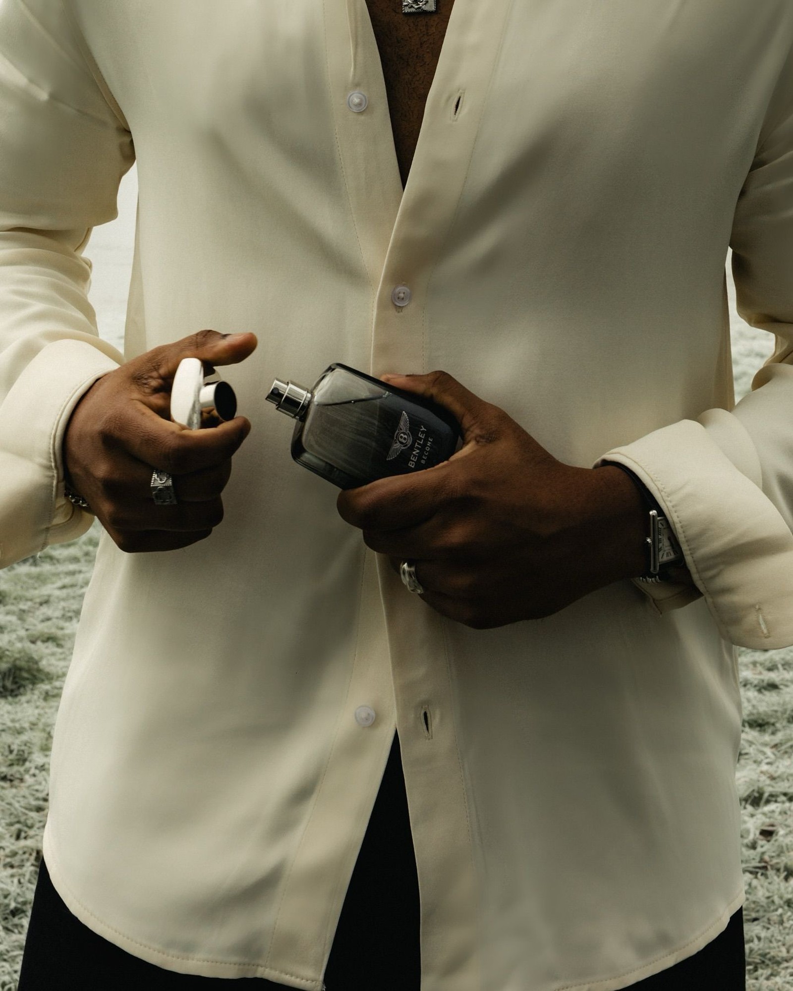 Man in a cream silk shirt holding a dark Bentley cologne bottle with the cap off in his other hand