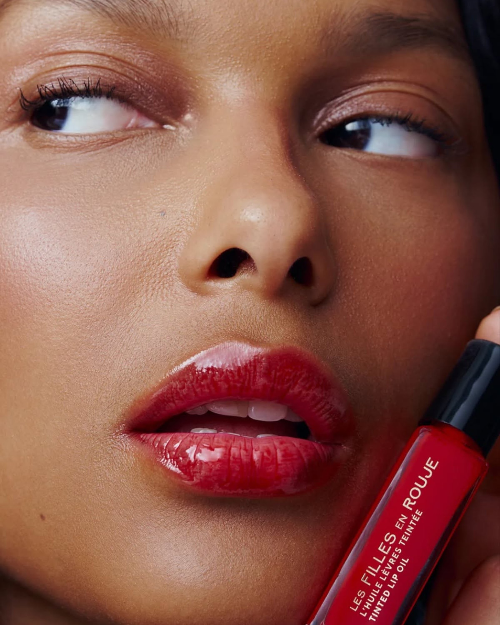 Close up of a woman's face with glossy red lips holding a red tinted lip oil tube