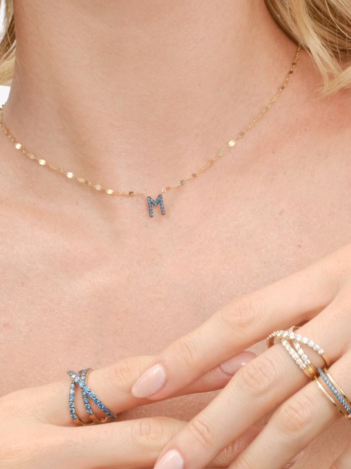 Close crop of a woman in a blue top wearing an initial necklace and stacked rings
