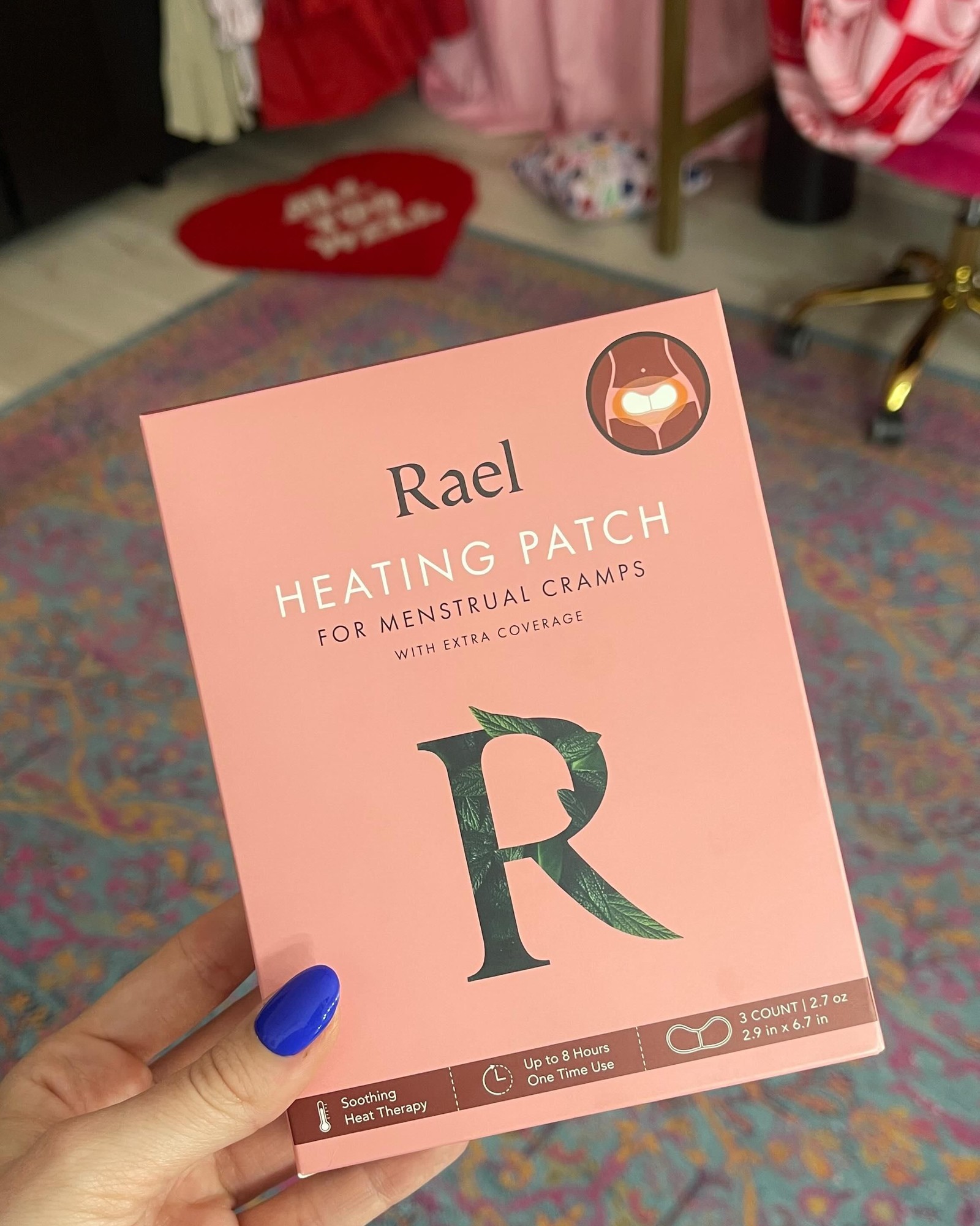 Hand with blue nails holding a coral pink box of Rael Heating Patch for Menstrual Cramps in a pink room