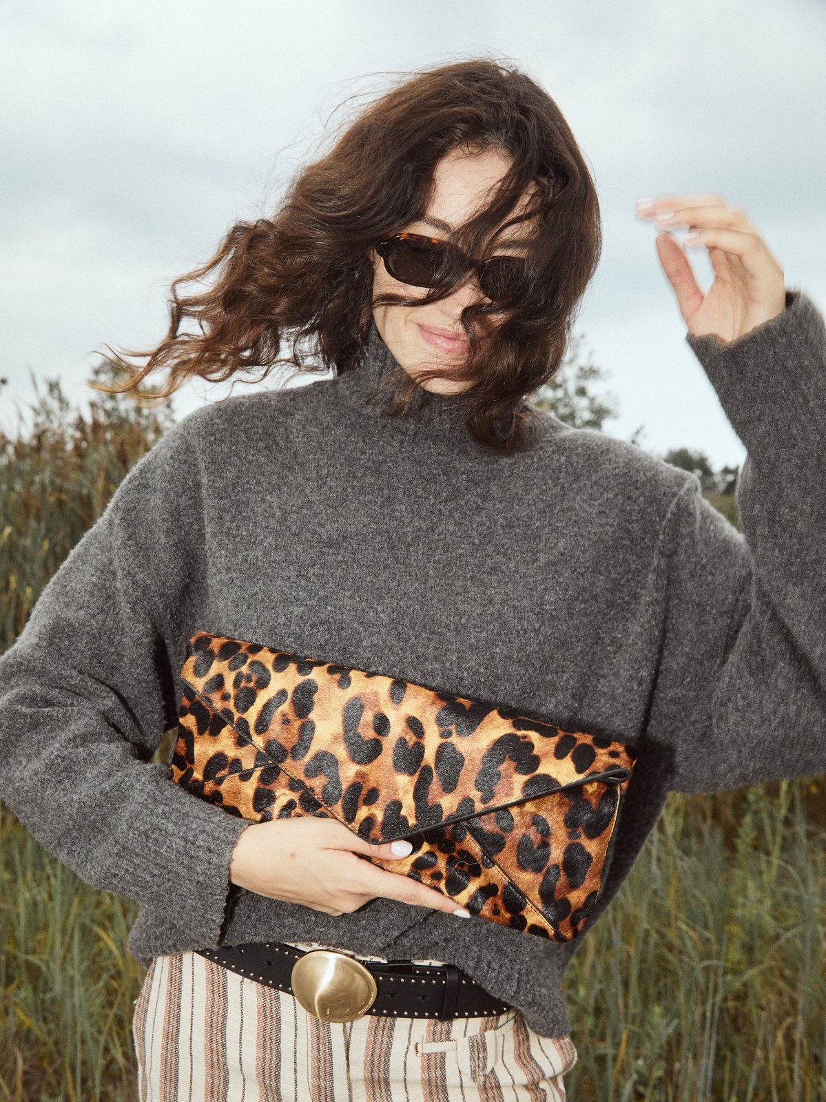 Model in a gray knit and striped trousers holding a leopard print clutch outdoors