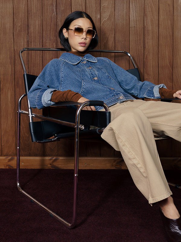 Model lounges in a denim jacket and tan trousers in a wood-paneled room