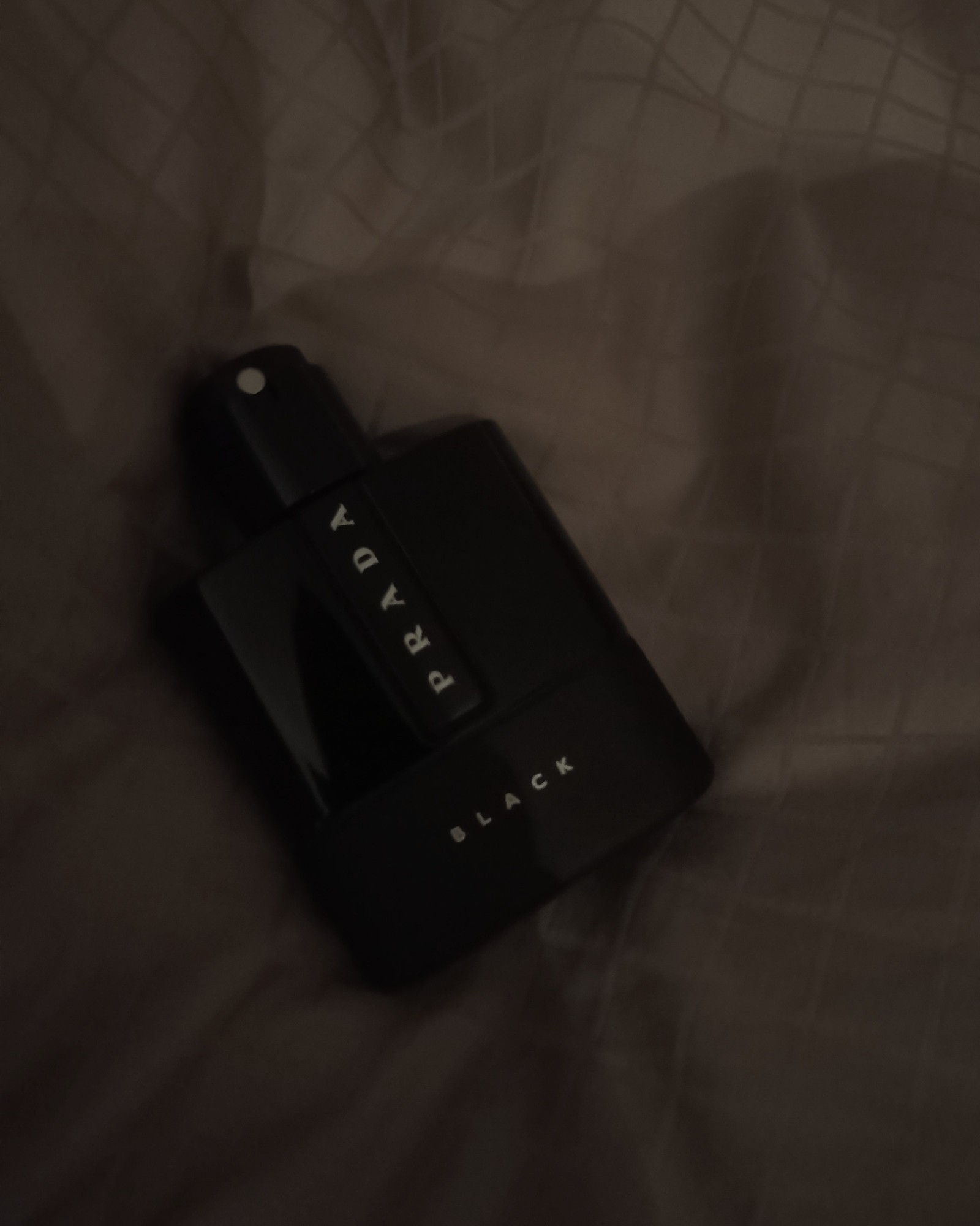 A dark, moody photo of a black Prada Luna Rossa Black bottle lying on creased bedsheets