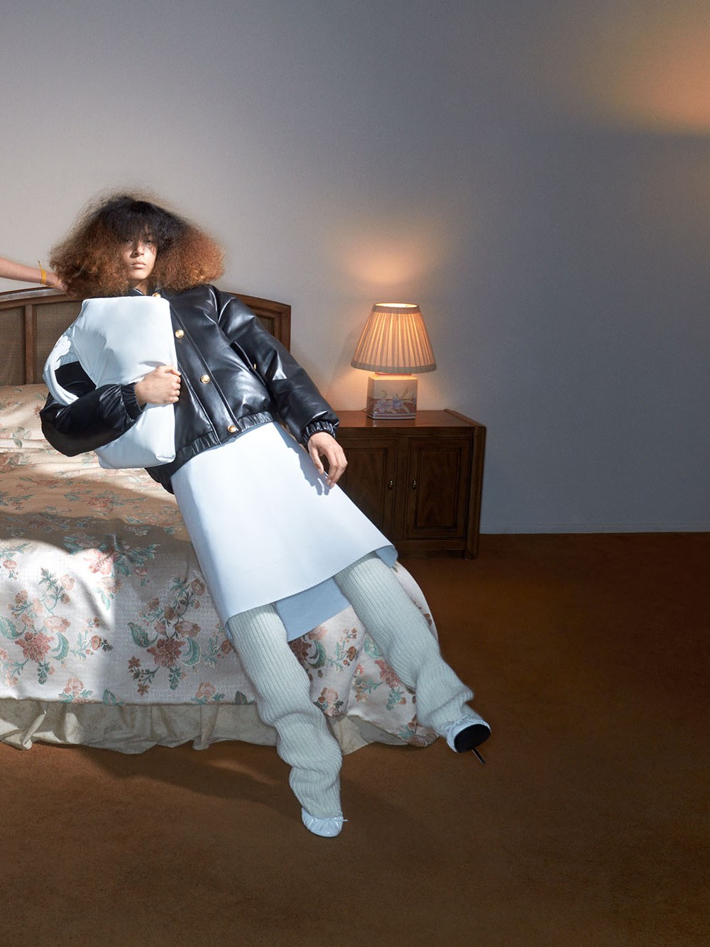 Model in a black jacket over a pale dress reclining on a bed