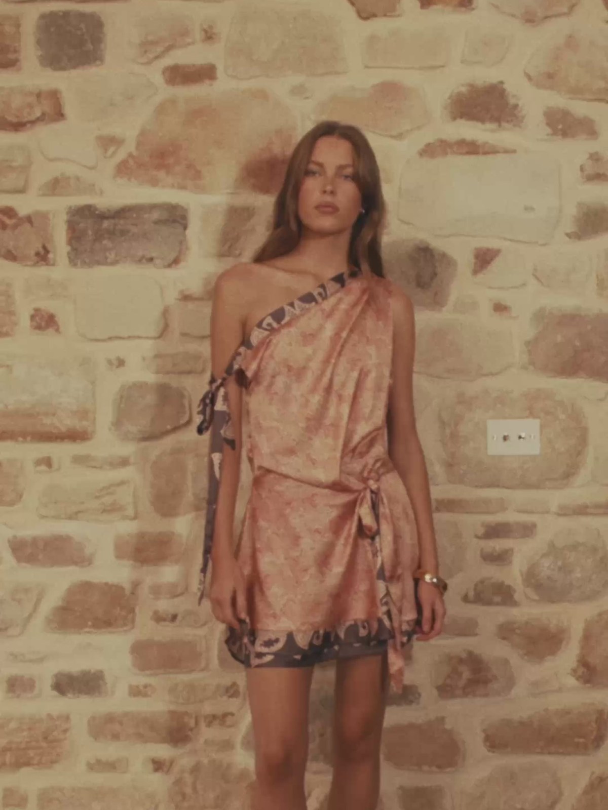 Model in a pink patterned mini dress against a stone wall