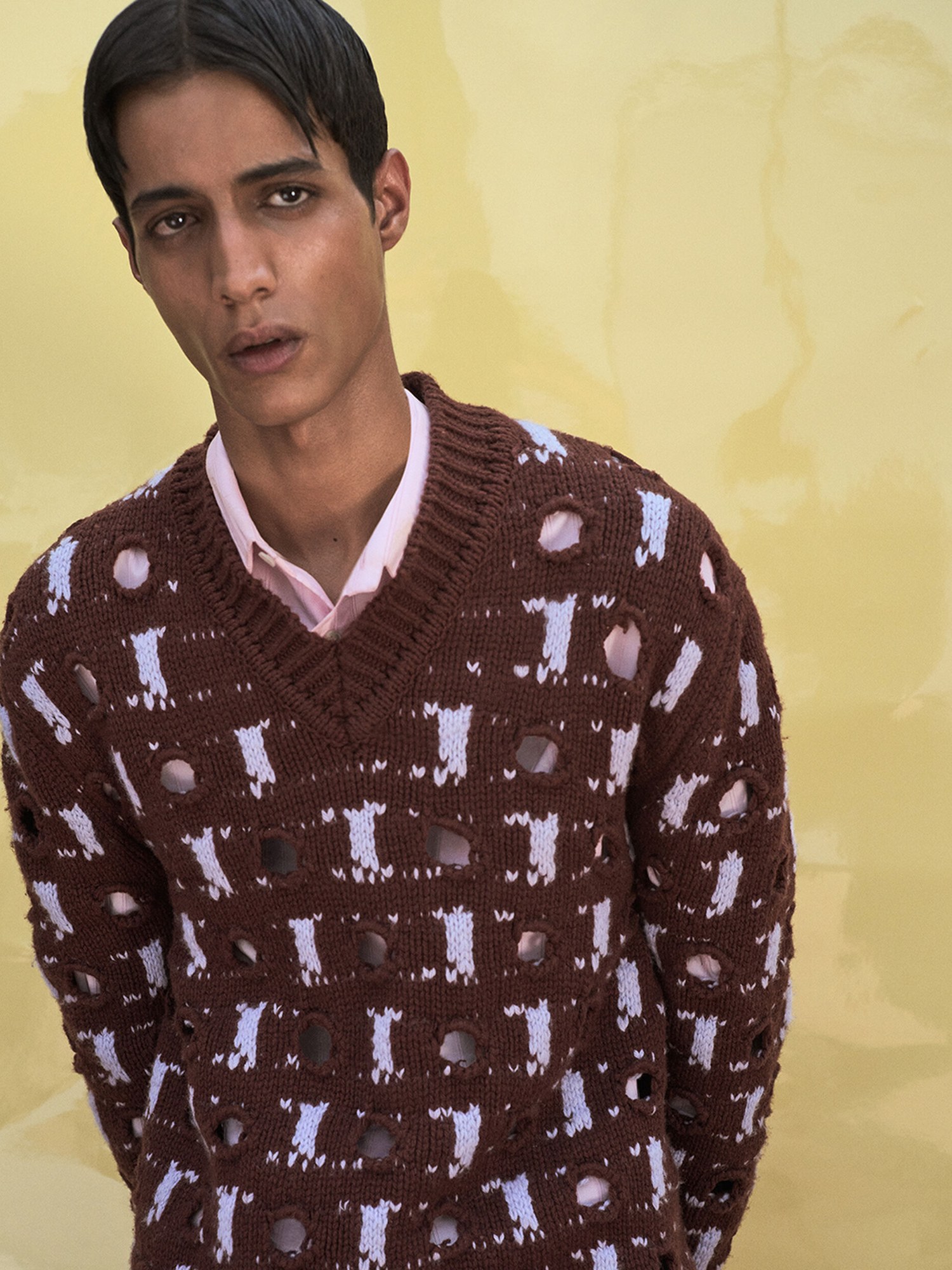 Model in a brown open knit sweater over a pink shirt against a yellow wall