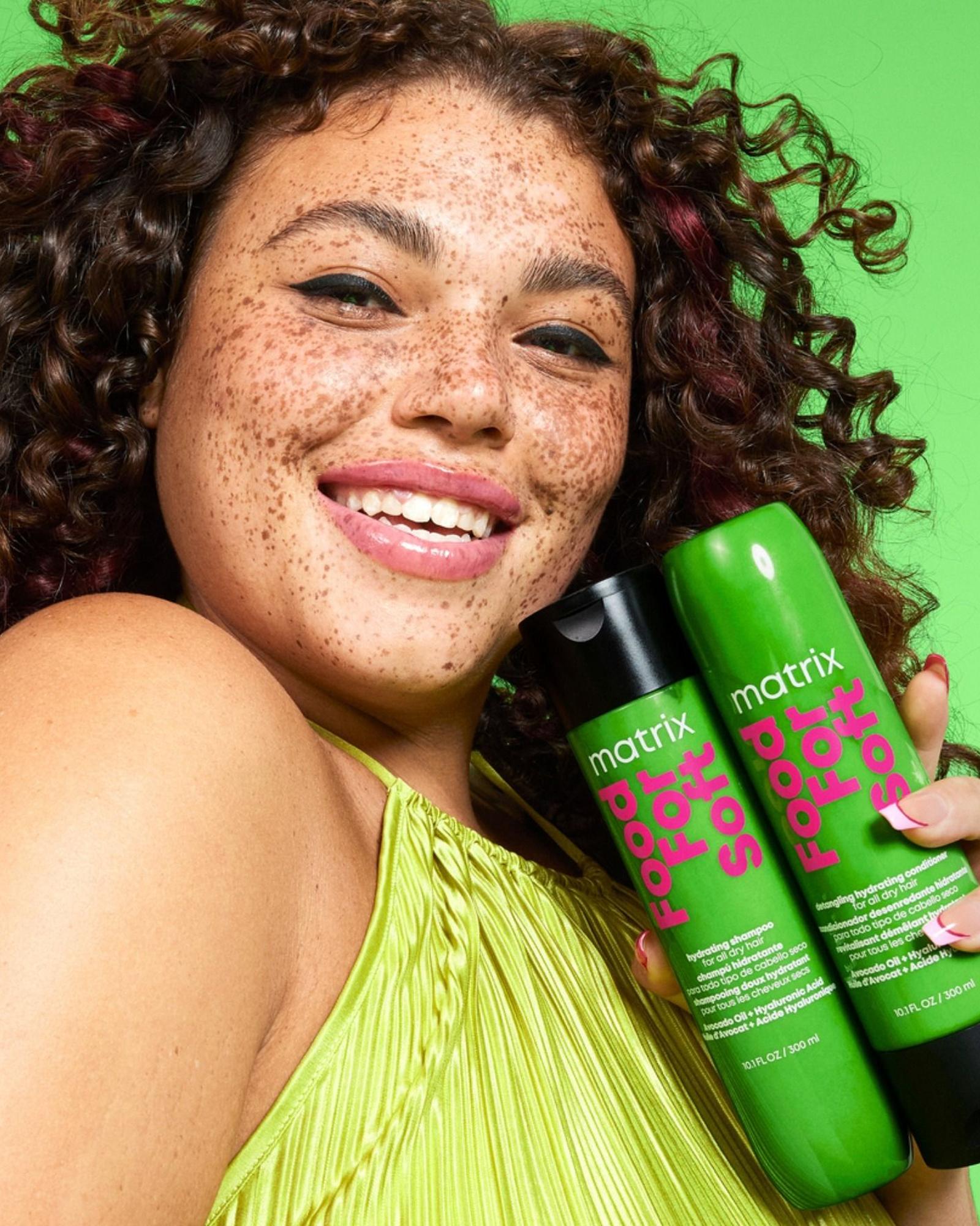 Smiling woman with freckles and dark curls holding green Matrix Food For Soft shampoo and conditioner bottles against a green background