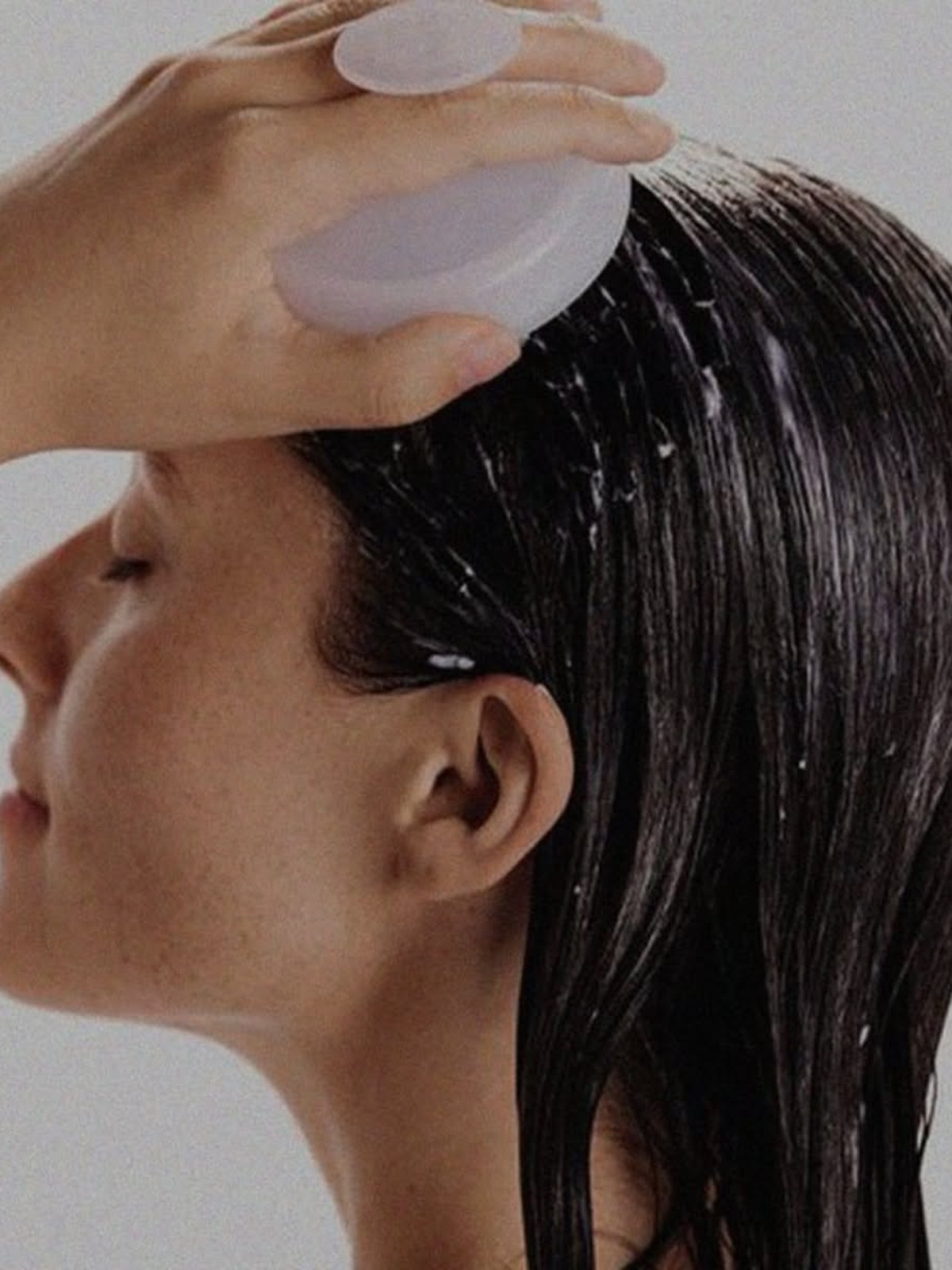 Side profile of a woman with wet dark hair using a pale lilac silicone scalp massager on her head, with conditioner visible in her hair