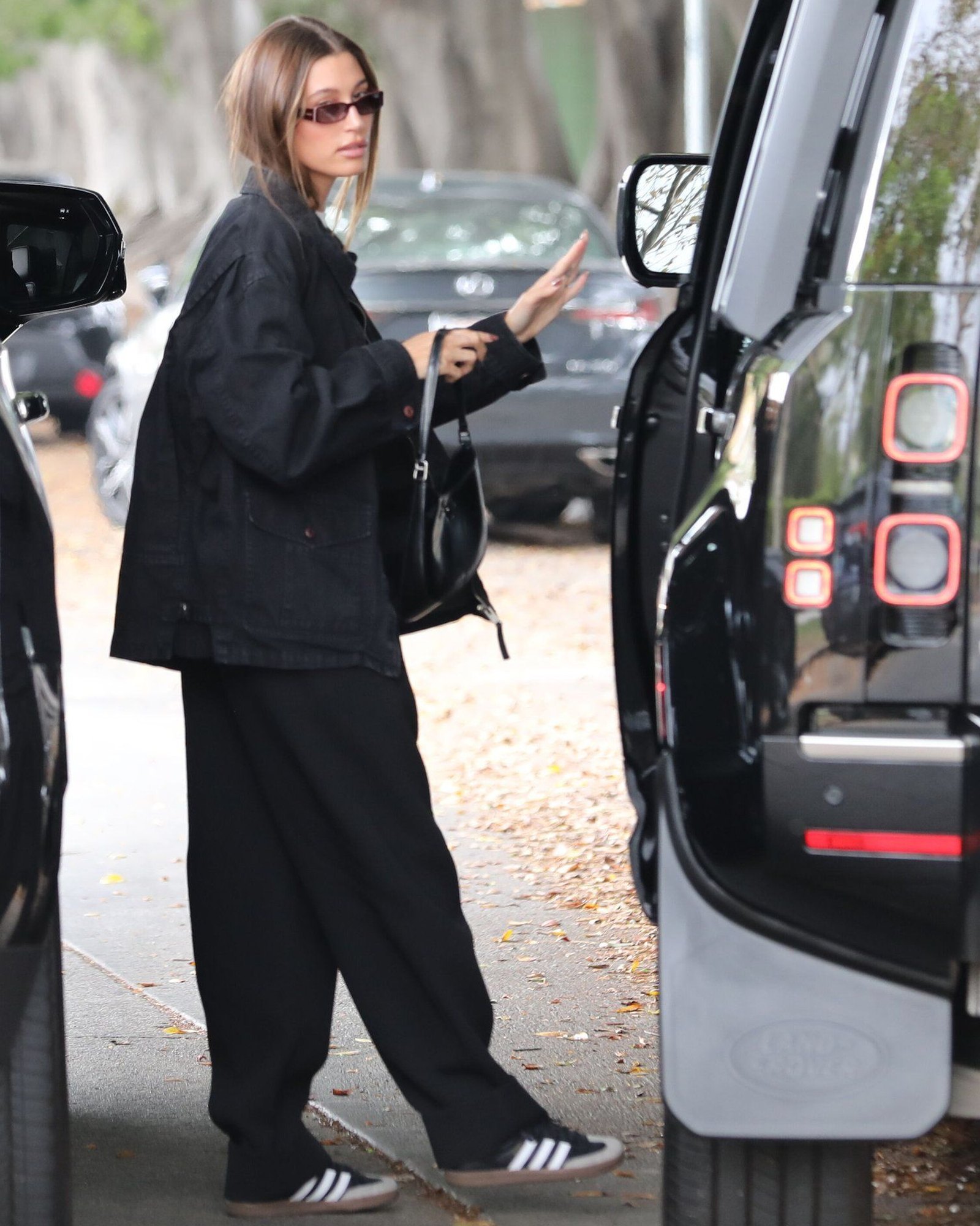 Woman on a street beside a car in a black jacket and black wide trousers with black and white adidas Samba sneakers