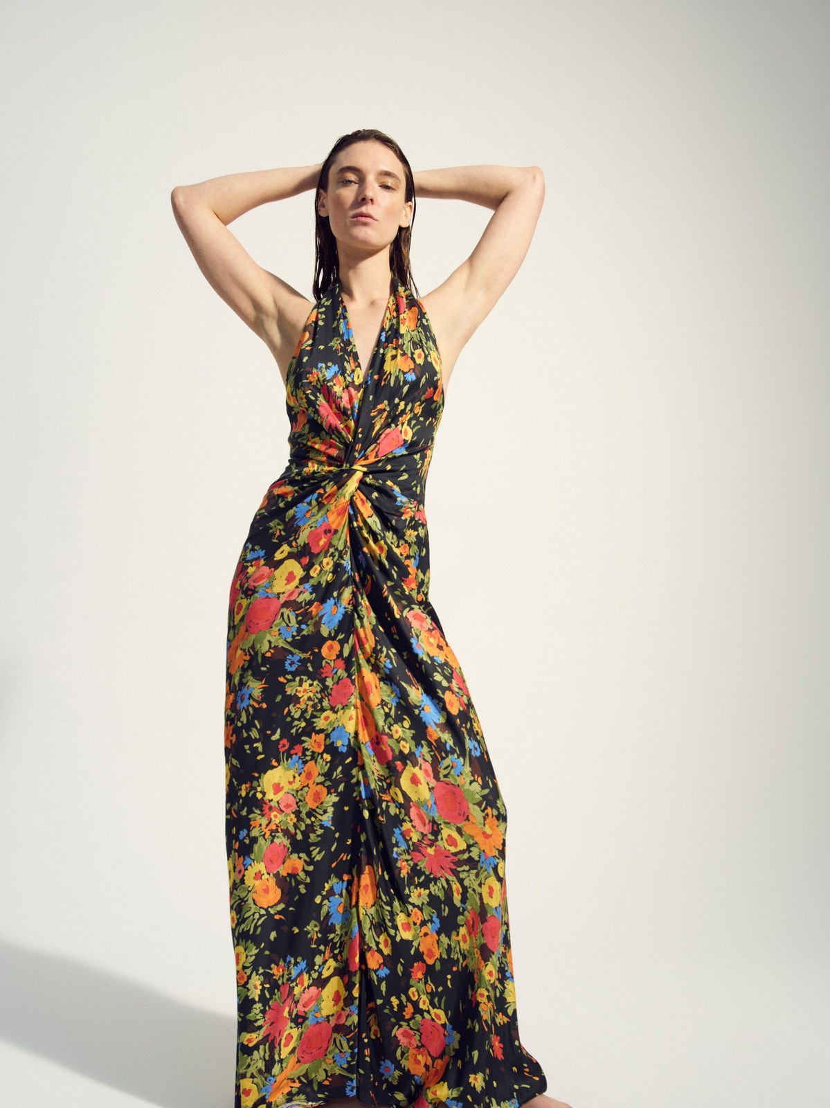 Model in a colorful floral gown with her arms raised against a pale studio backdrop