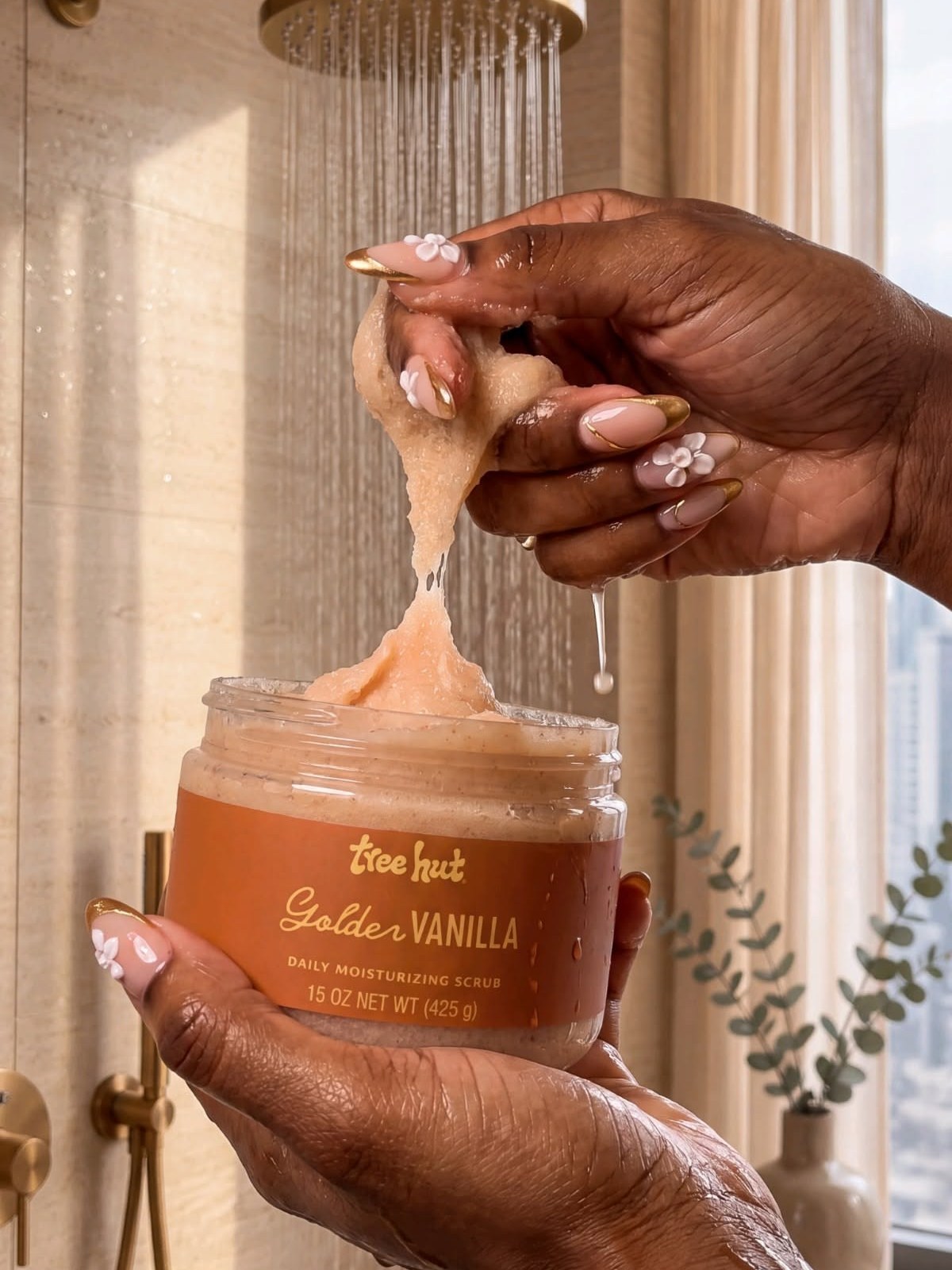 Hands with pale floral nail art holding a jar of Tree Hut Golden Vanilla scrub, scooping some out in front of a gold rain shower head