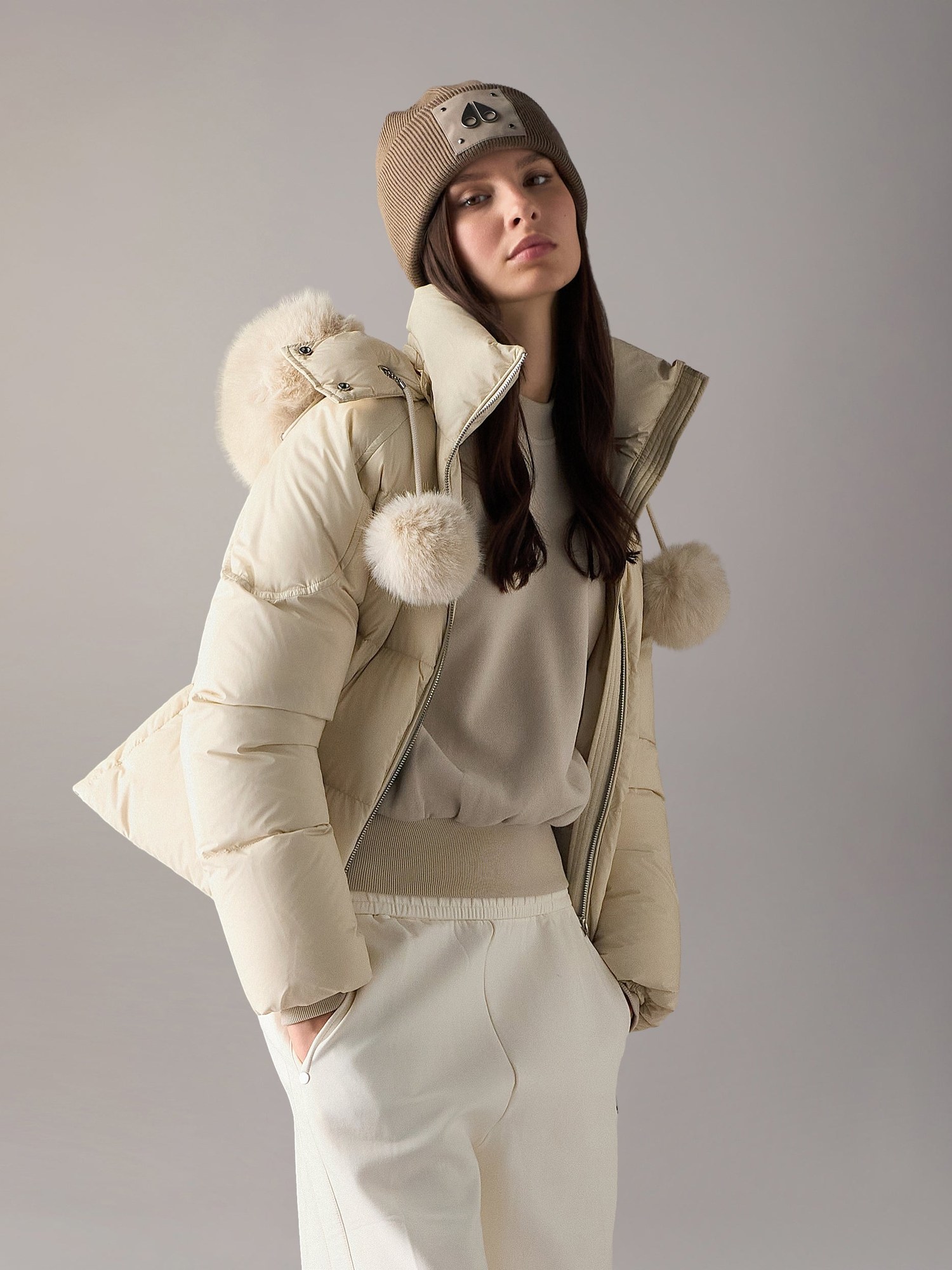 Model in a cream puffer jacket with pom pom fur trim and a beanie