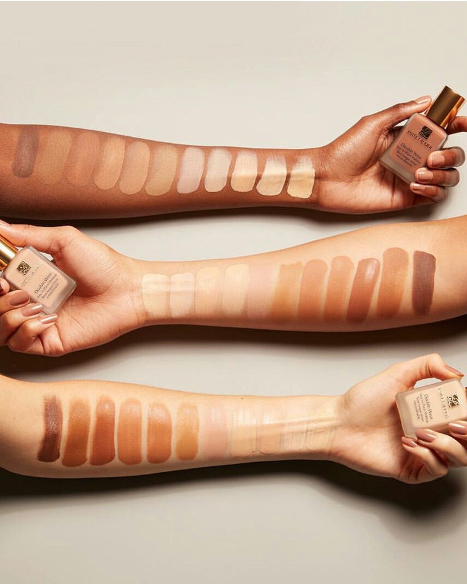 Three forearms with rows of Double Wear foundation swatches in light to deep shades, each hand holding a bottle, on a beige background