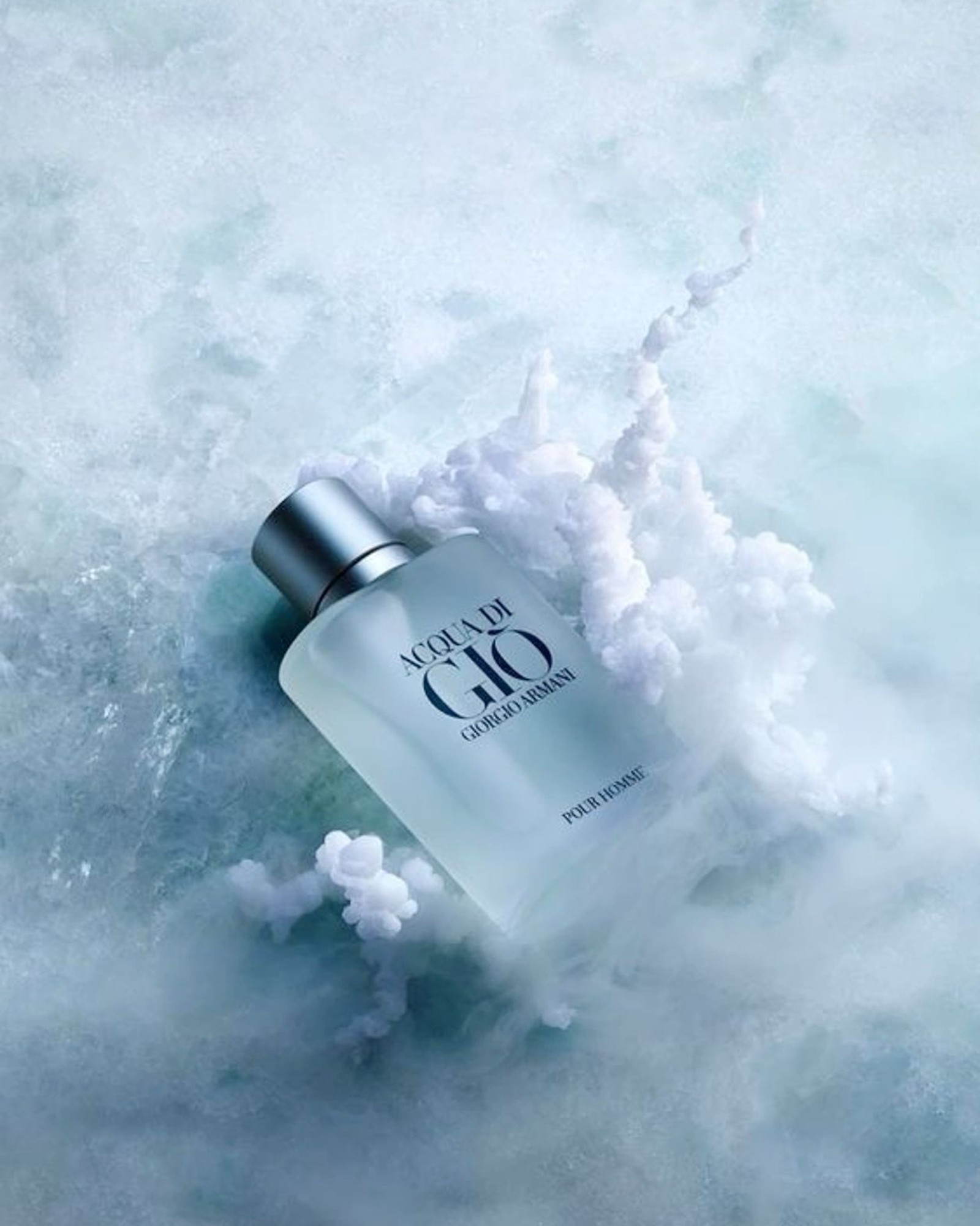 Frosted bottle of Acqua di Giò Pour Homme lying on ice with swirls of white cloud rising beside it in pale blue-green tones.