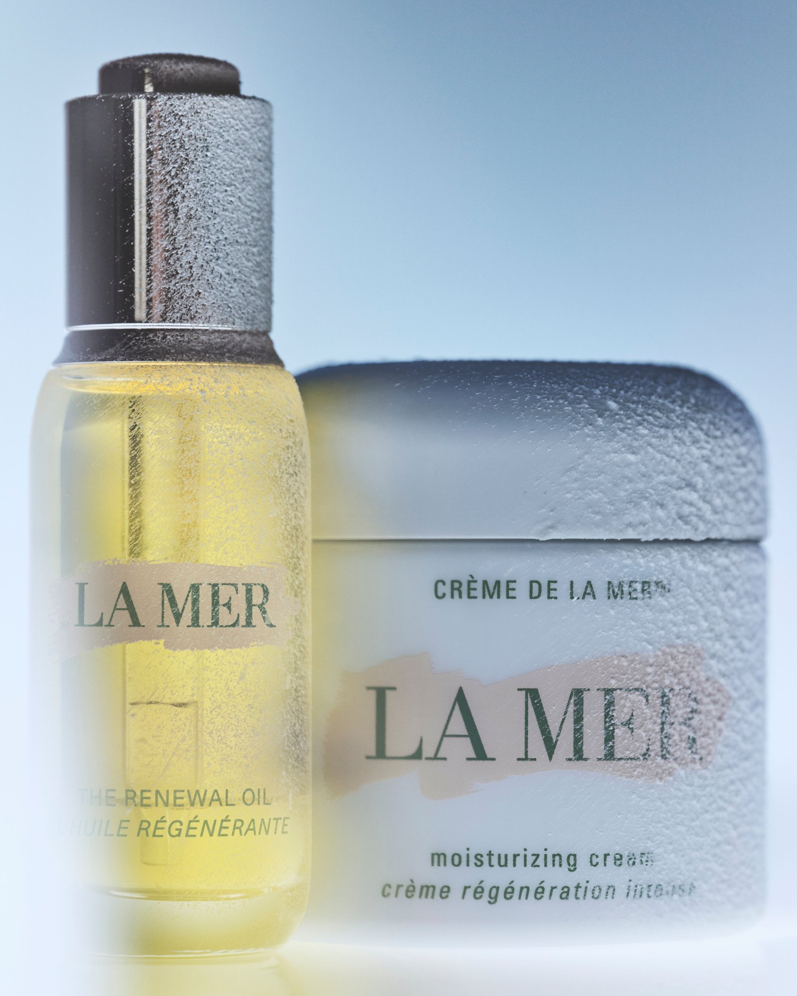 La Mer The Renewal Oil dropper bottle beside a Crème de la Mer jar, both dusted with frost against a pale blue background