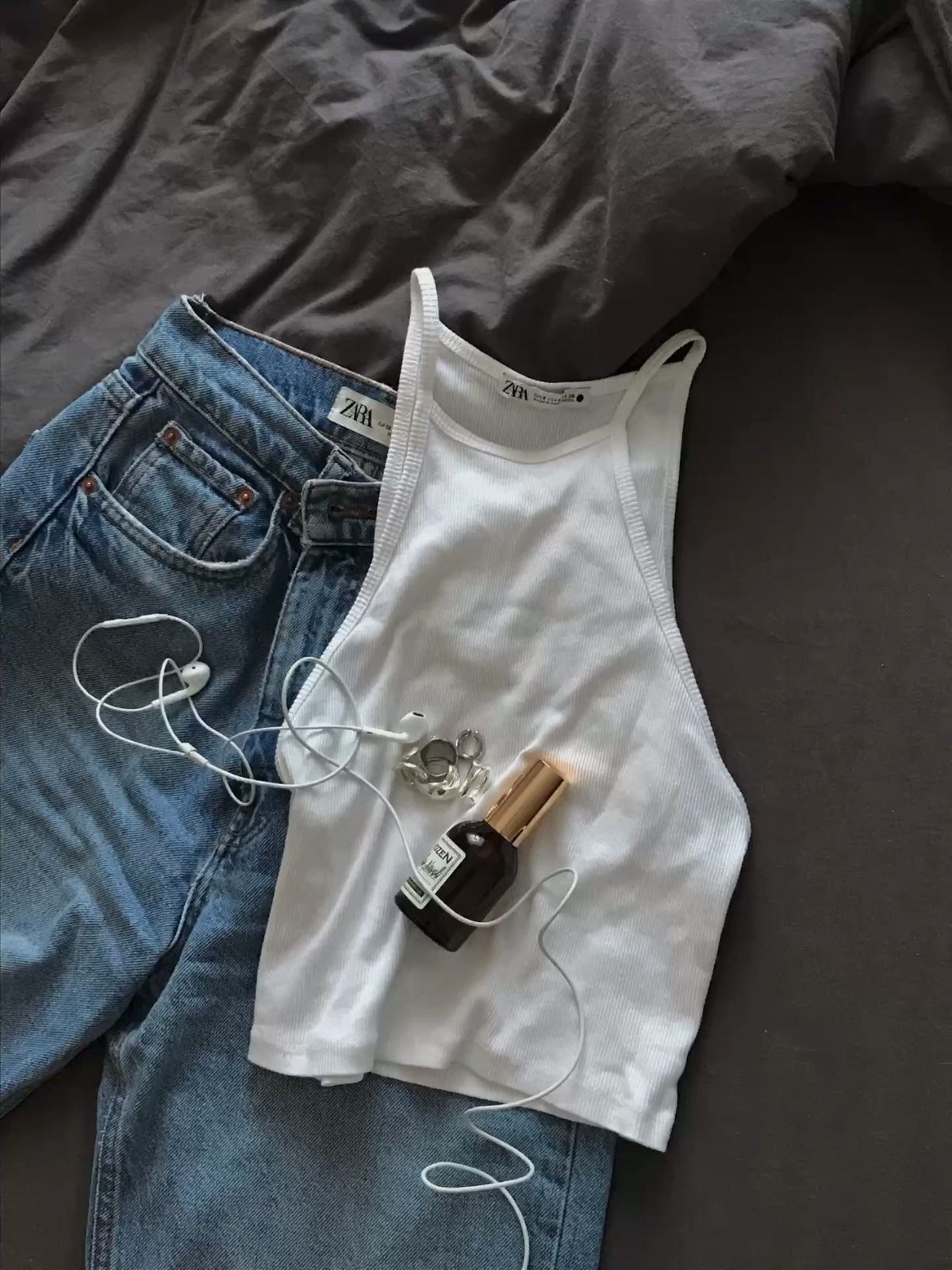 Jeans and a white tank laid out on a bed with headphones and perfume