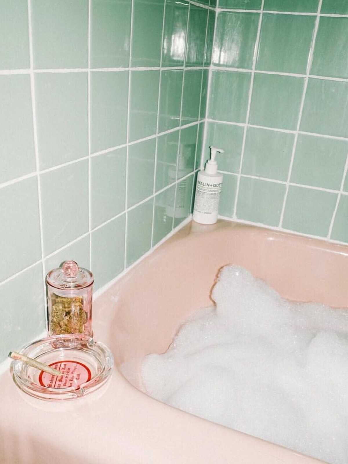 Pink bathtub full of white bubbles against mint green tiles, with a white pump bottle on the ledge