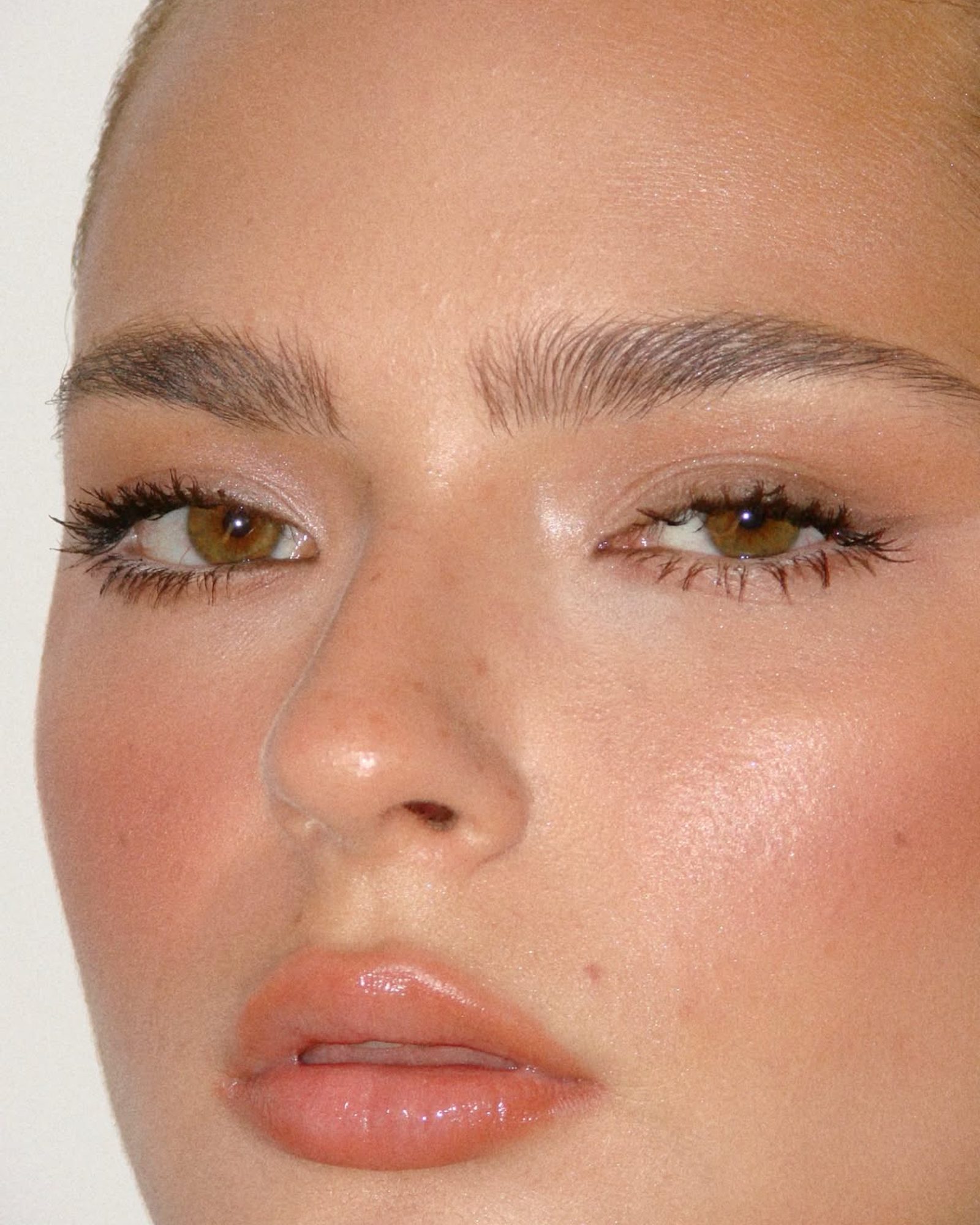 Close-up of a woman with hazel eyes, soft brown lashes, a glossy peach lip and a dewy flushed cheek