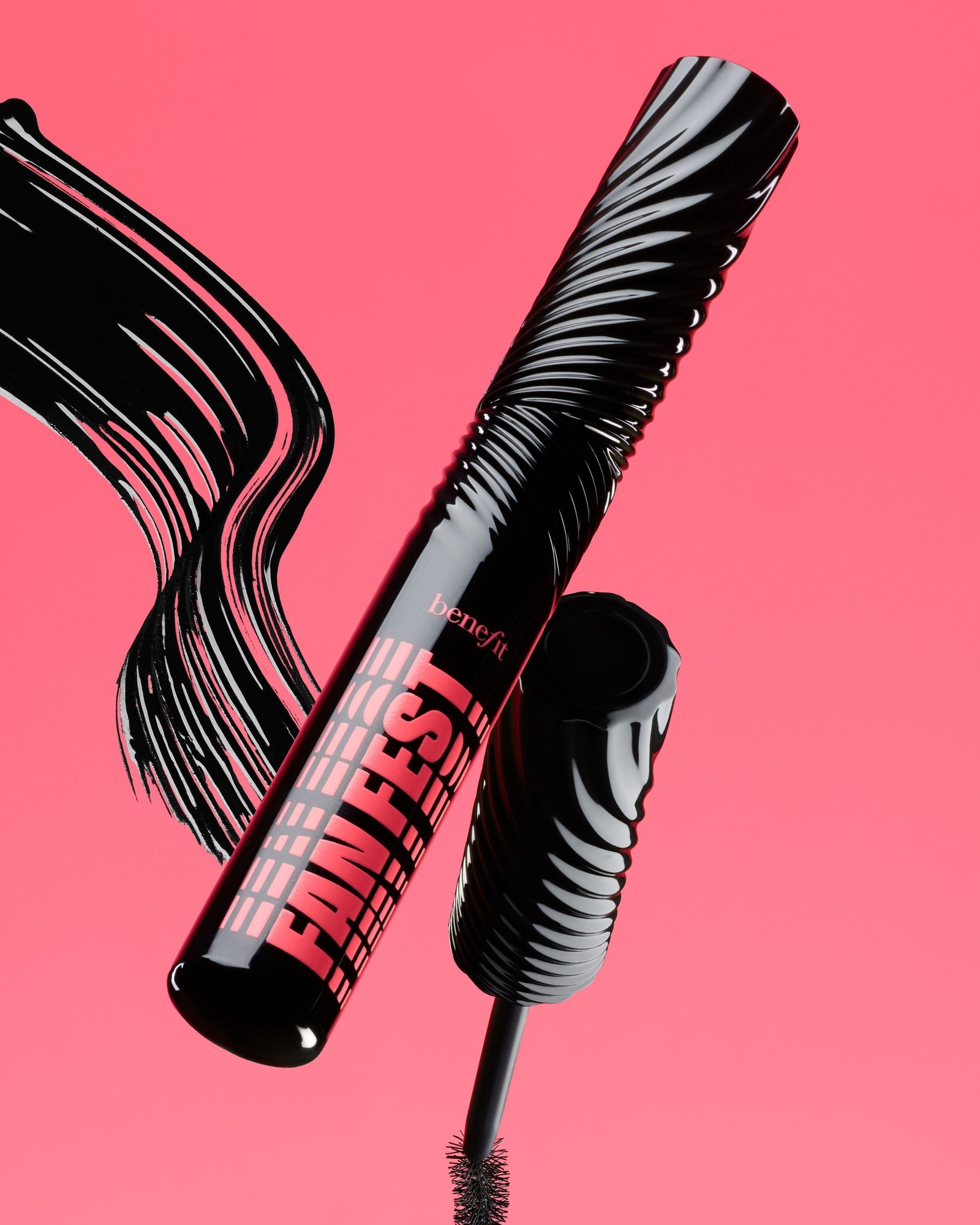 Close-up of a black Benefit Fan Fest mascara tube and its wand tip beside a thick black brush stroke on a coral pink background