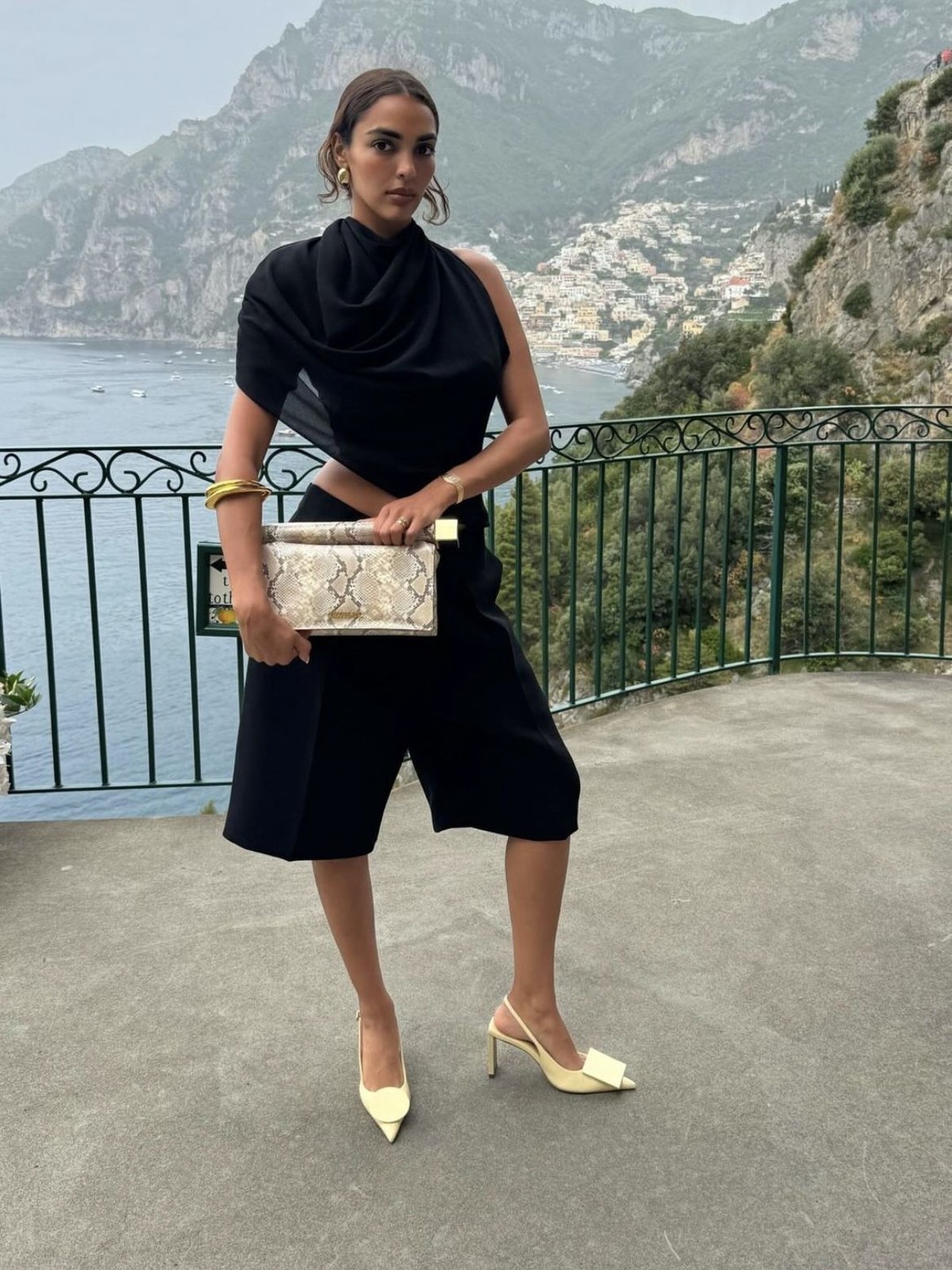 Woman in a black draped top and shorts with pale heels and a snakeskin clutch on a terrace above the sea