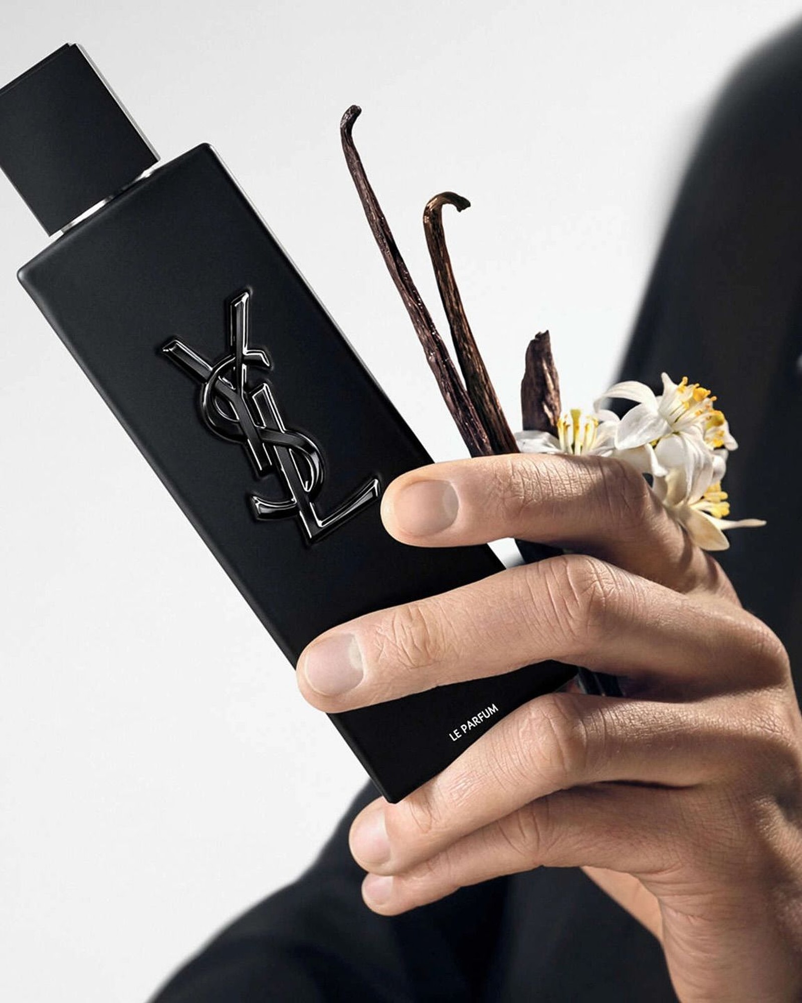 Man's hand holding a black YSL MYSLF Le Parfum bottle with vanilla pods and white orange blossom flowers