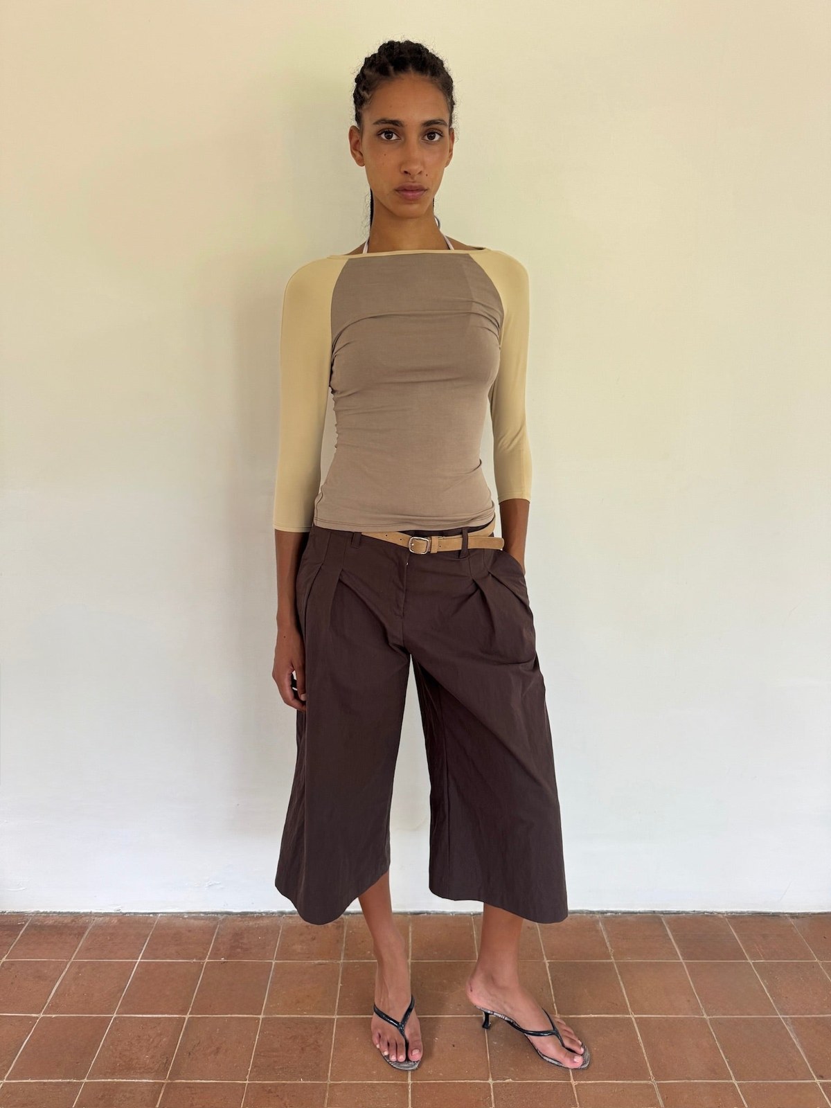 Model wearing a beige top and brown cropped trousers against a warm tiled wall
