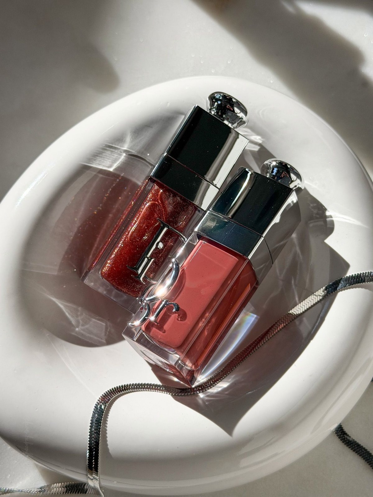 Two Dior Addict lip glosses, one glittery red and one soft rose, in a white dish with a silver snake chain