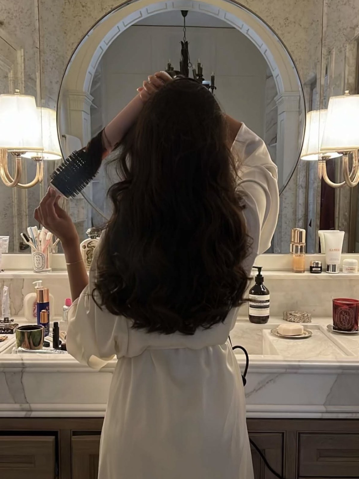 Woman in a white robe seen from behind holding a round brush in long dark wavy hair at a marble vanity with a mirror