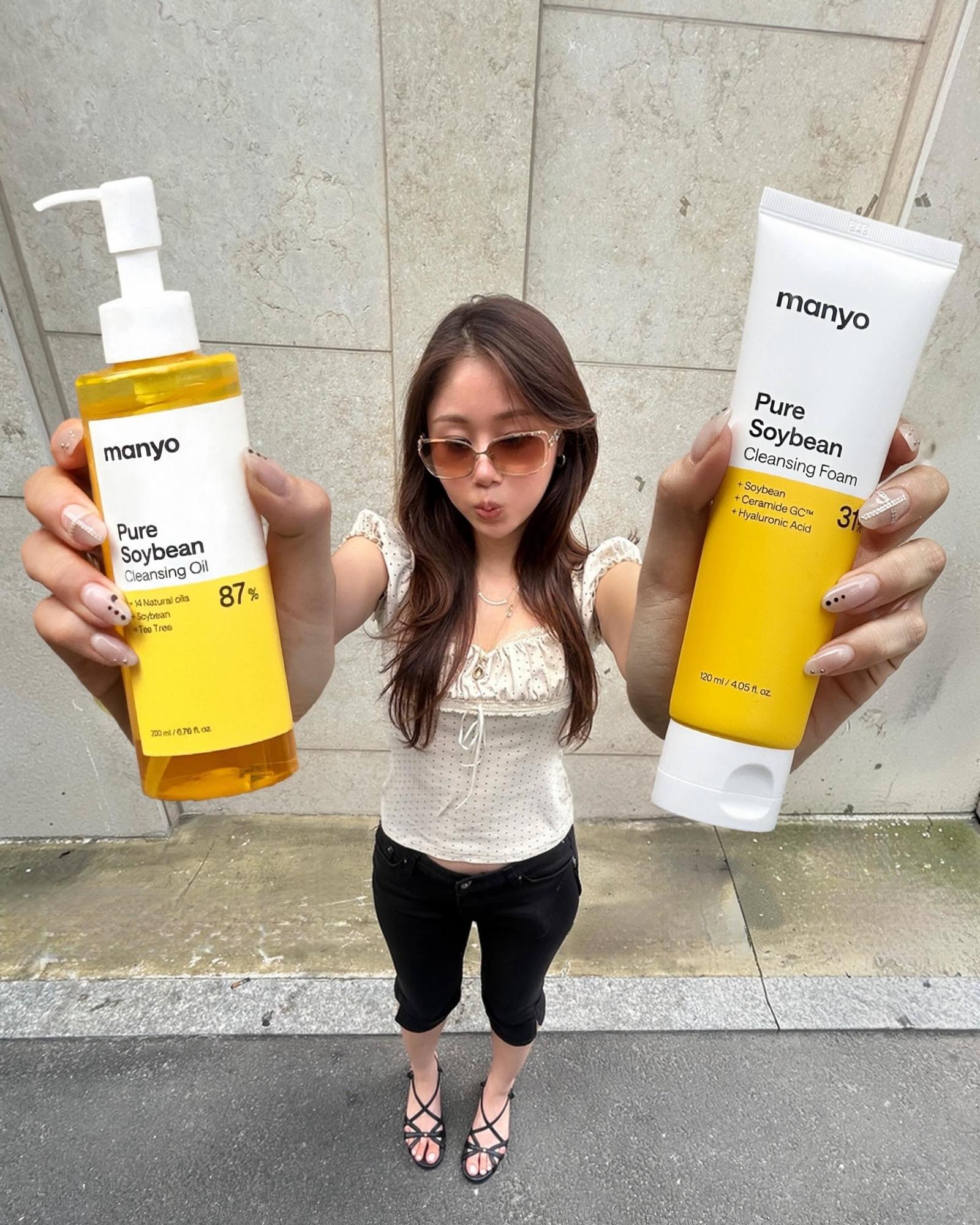Woman in tinted sunglasses holds up a bottle of manyo Pure Soybean Cleansing Oil in one hand and a tube of Pure Soybean Cleansing Foam in the other.