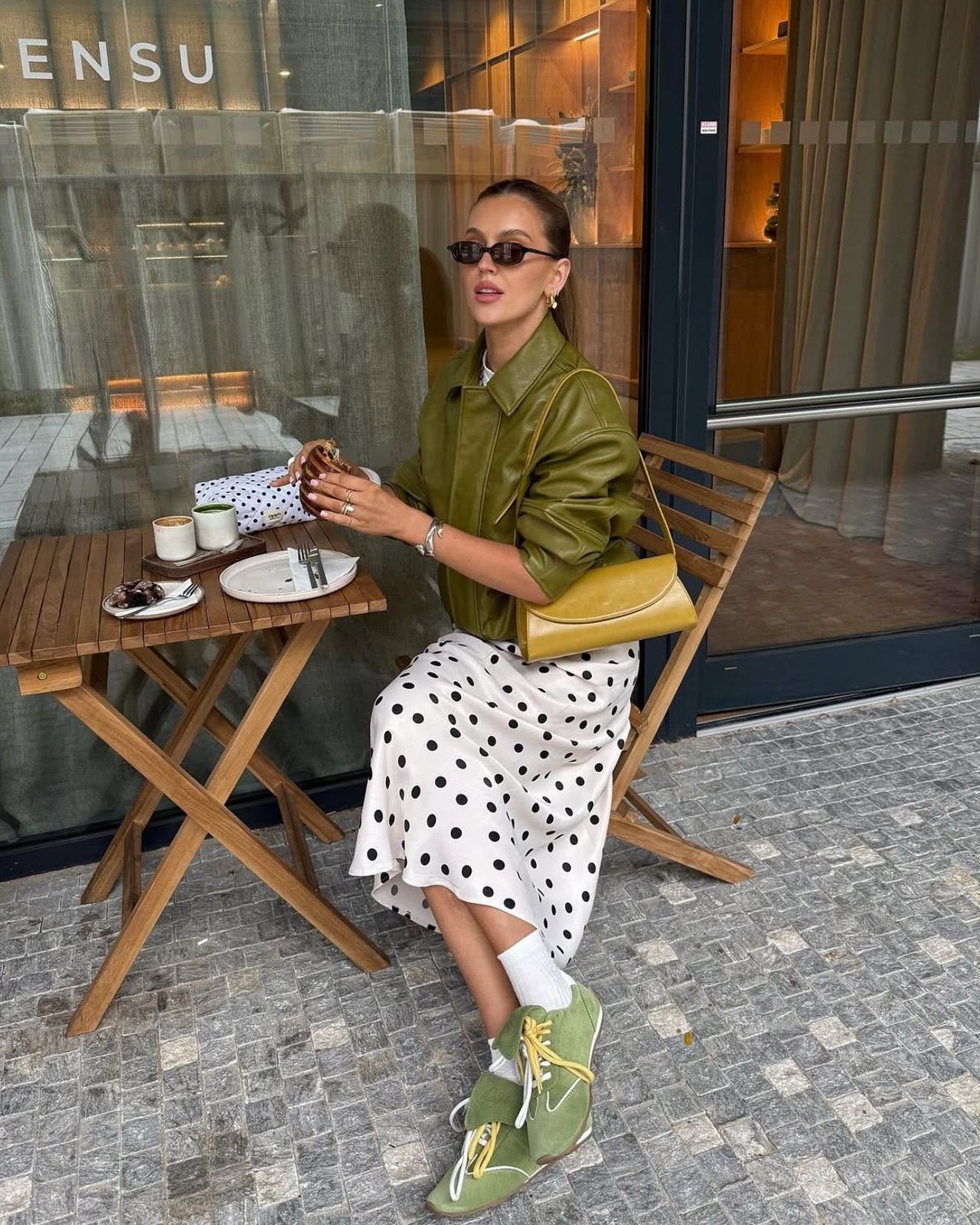 Woman at a cafe table in an olive leather jacket and a white midi skirt with black polka dots, wearing green sneakers