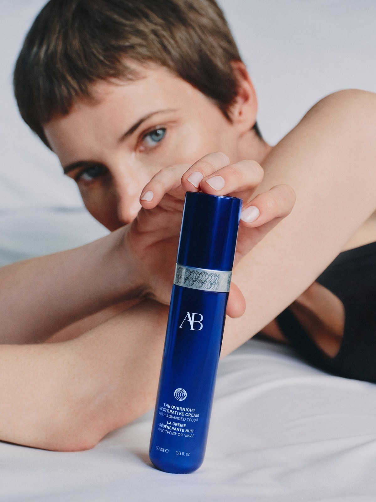 Short-haired woman reclining on pale bedding with an overnight cream bottle