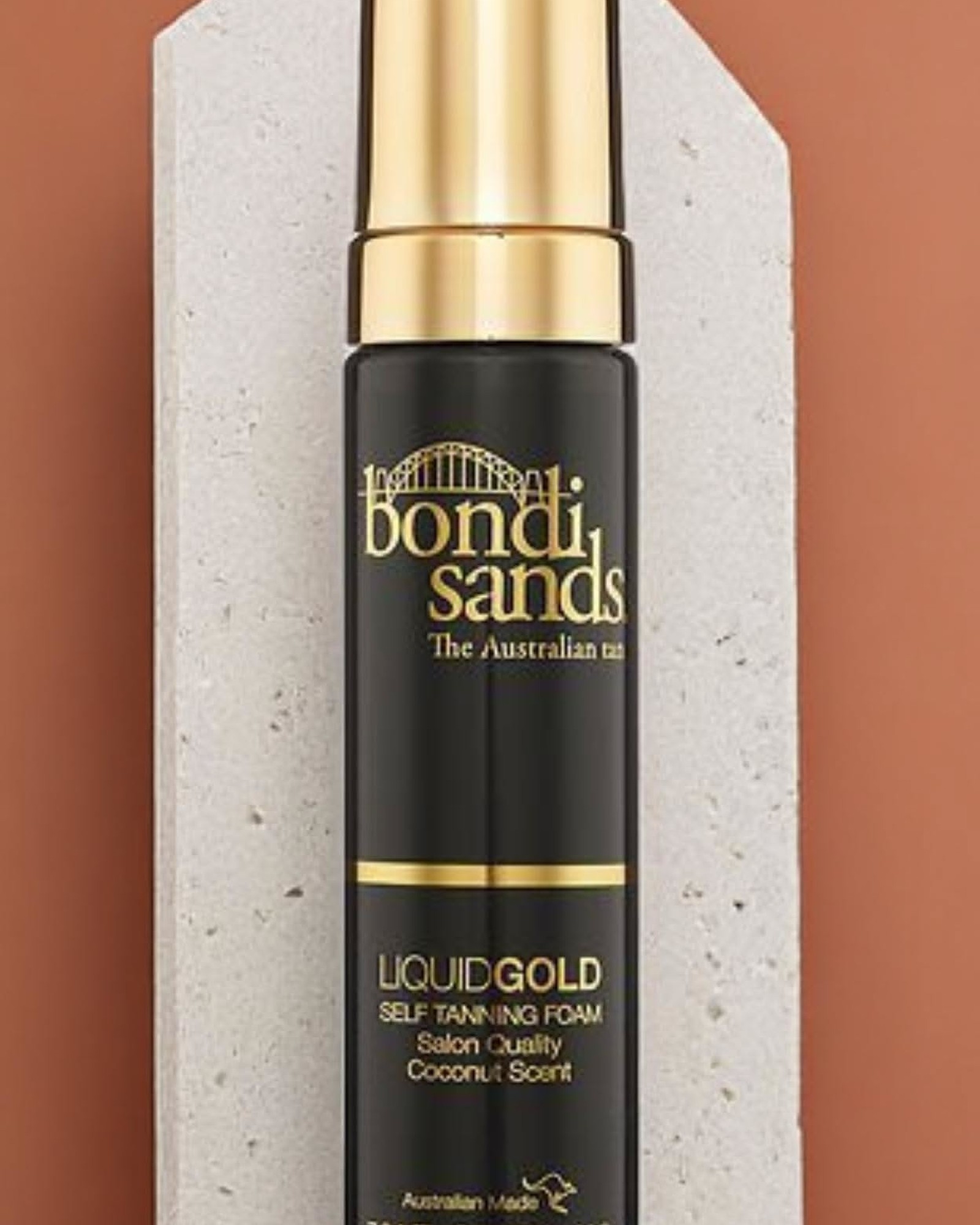 Bondi Sands Liquid Gold Self Tanning Foam in a black and gold bottle standing on a pale stone block against a terracotta background