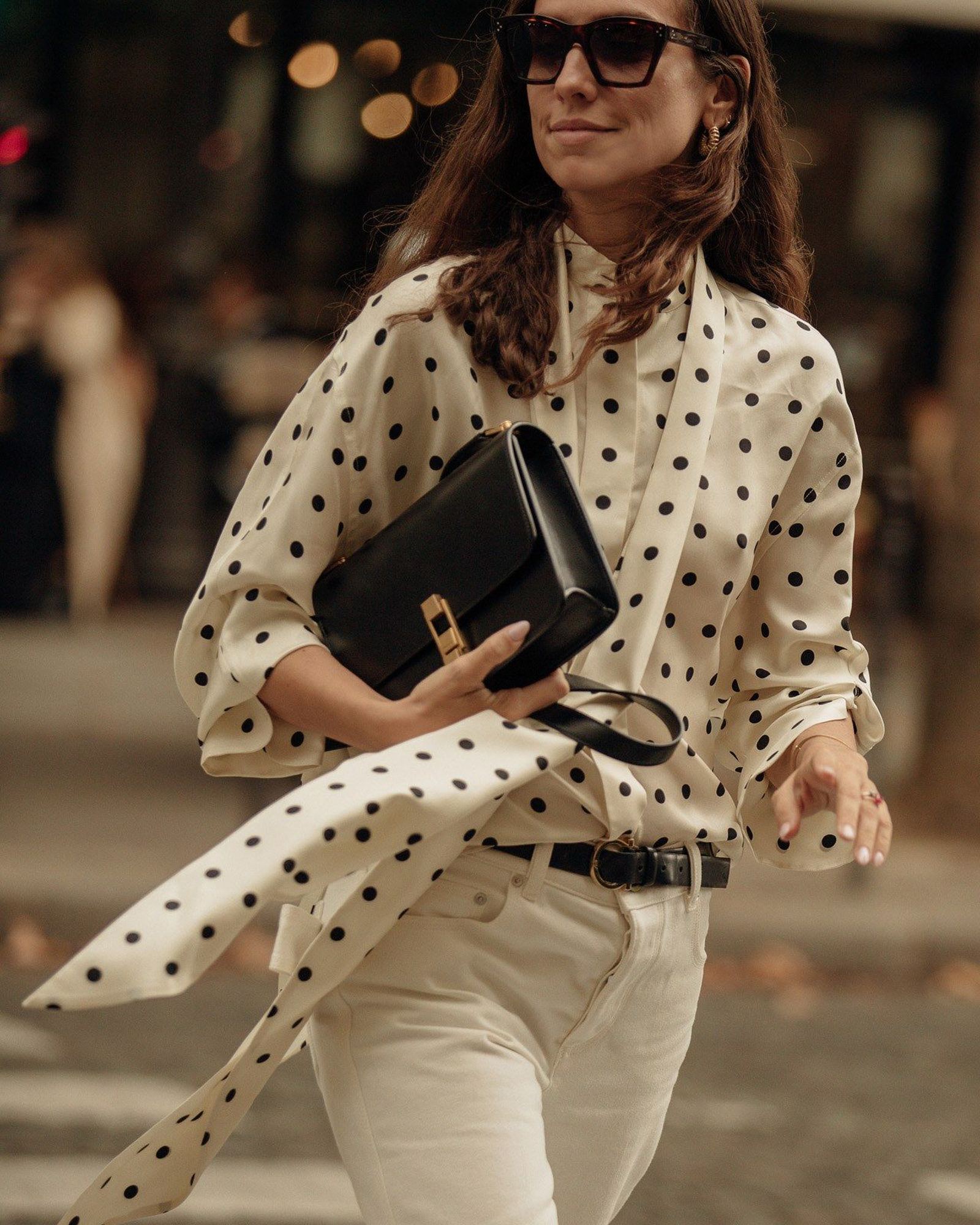 Woman in a cream polka dot blouse with a long scarf neck tucked into white trousers, holding a black shoulder bag