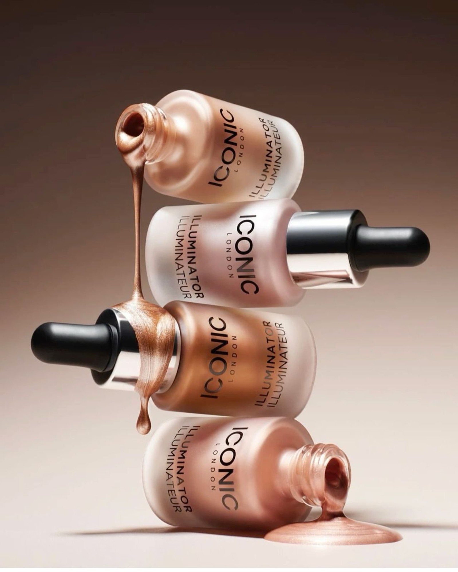 Four frosted bottles of ICONIC London Illuminator drops stacked on their sides, with shimmering bronze and pink liquid pouring from two of them