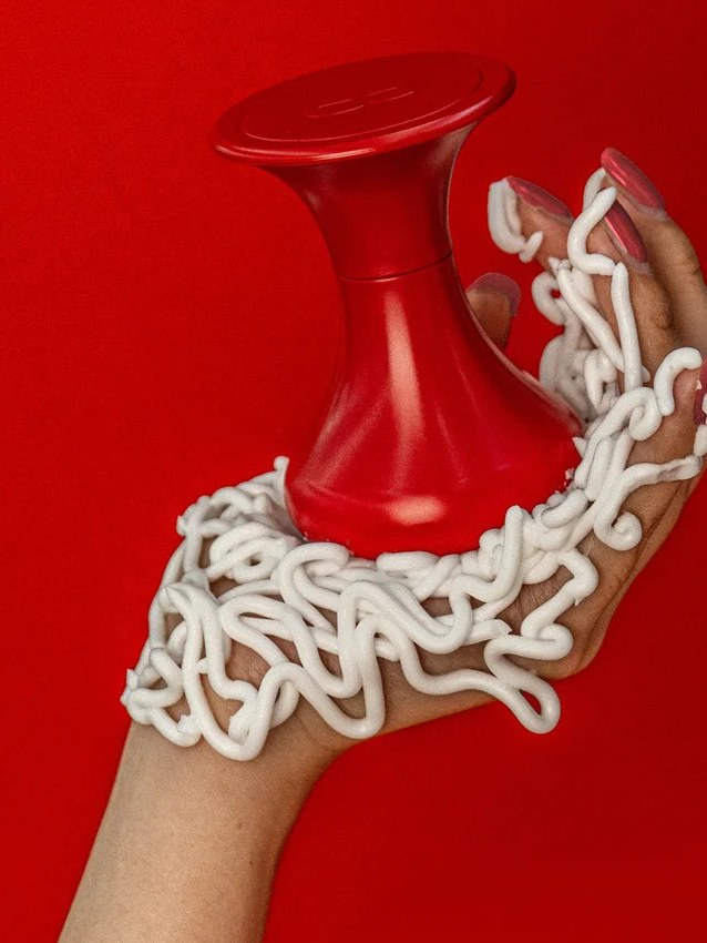 A hand coated with white cream curls around a red bottle against scarlet.