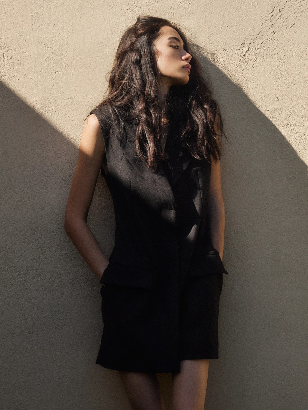 Model in a sleeveless black outfit against a sunlit stucco wall