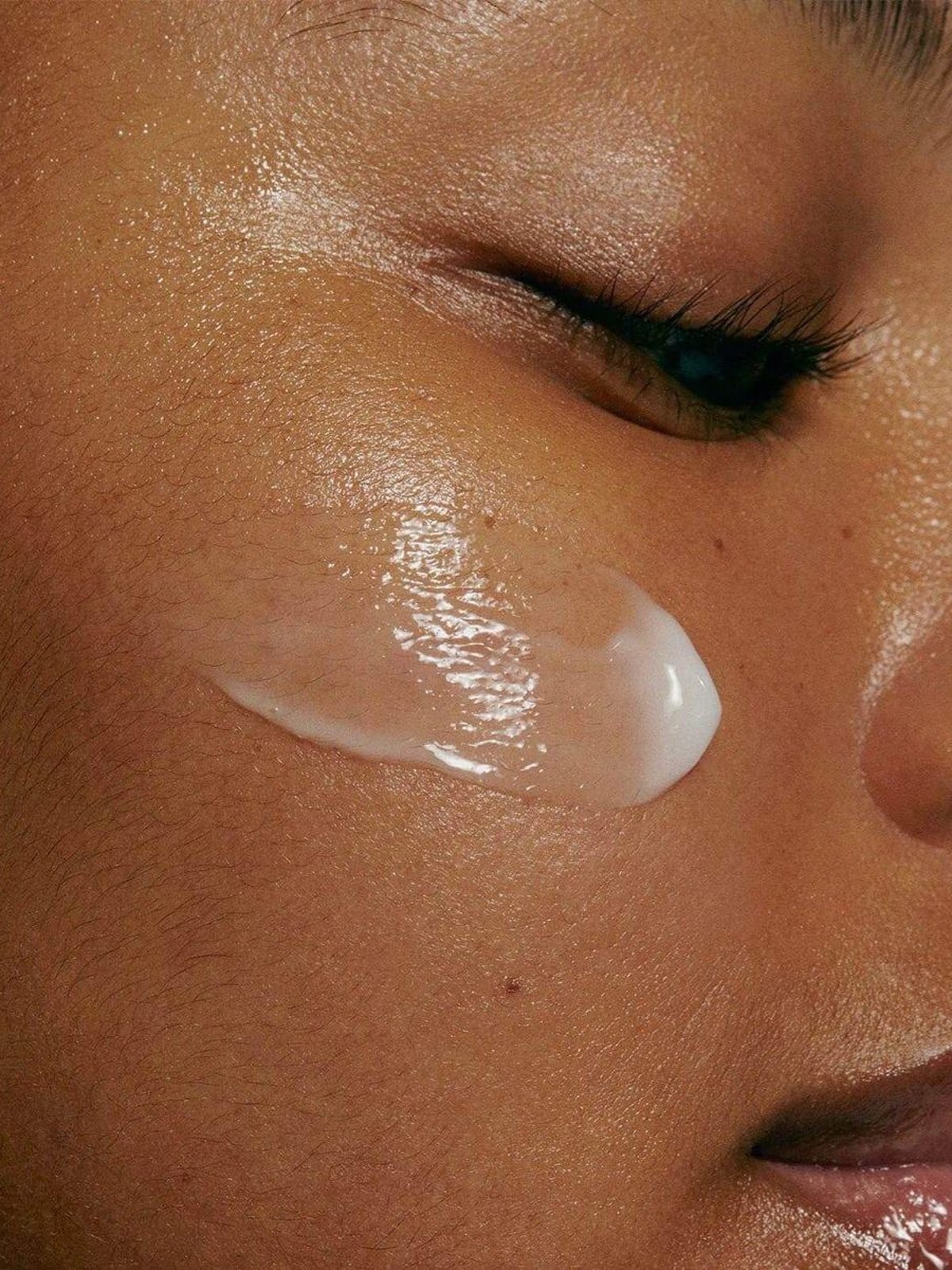 Close-up of a glossy white swipe of moisturizer on a woman's cheek beside her closed eye