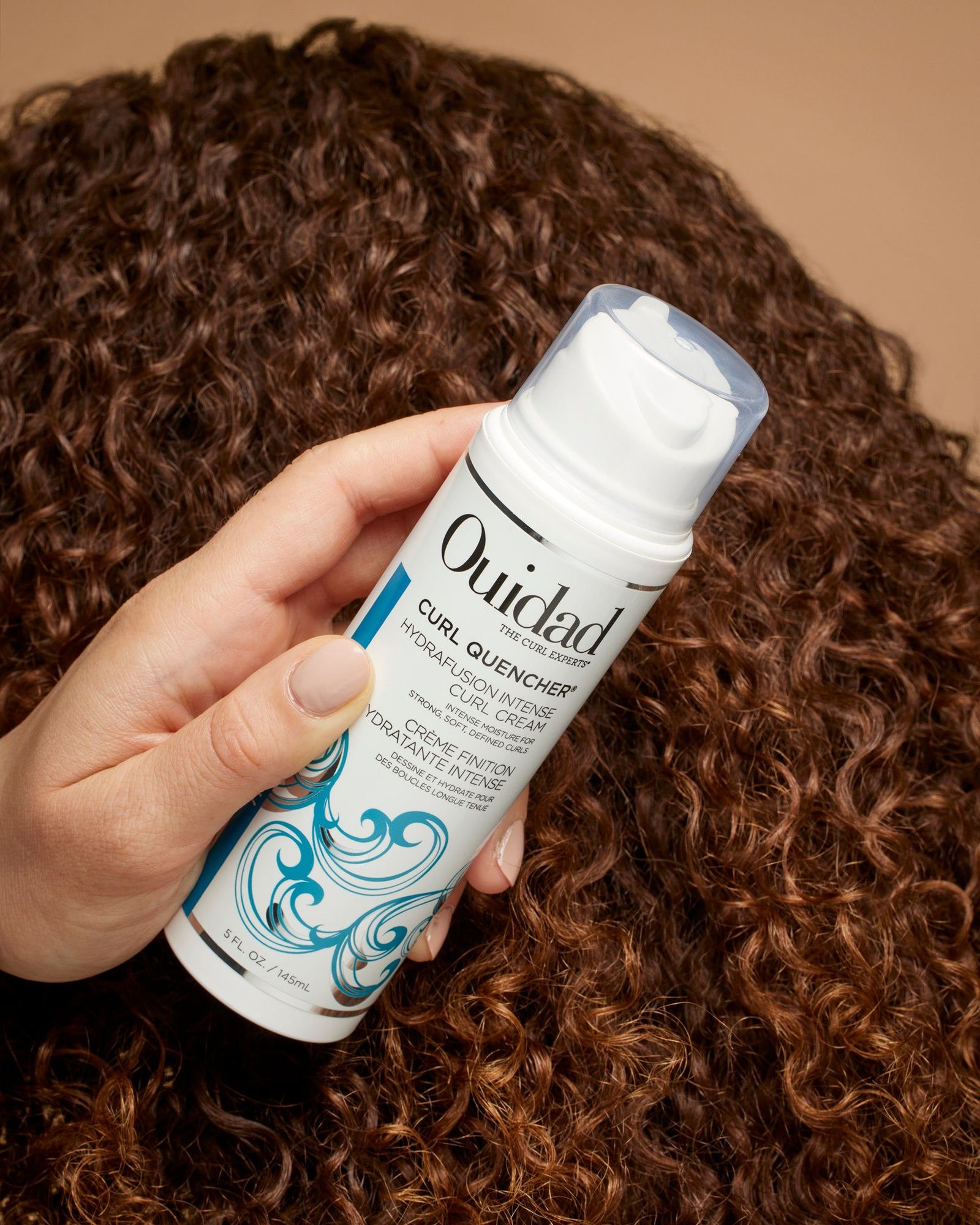 Hand holding a Curl Quencher Hydrafusion Intense Curl Cream pump bottle over auburn curly hair