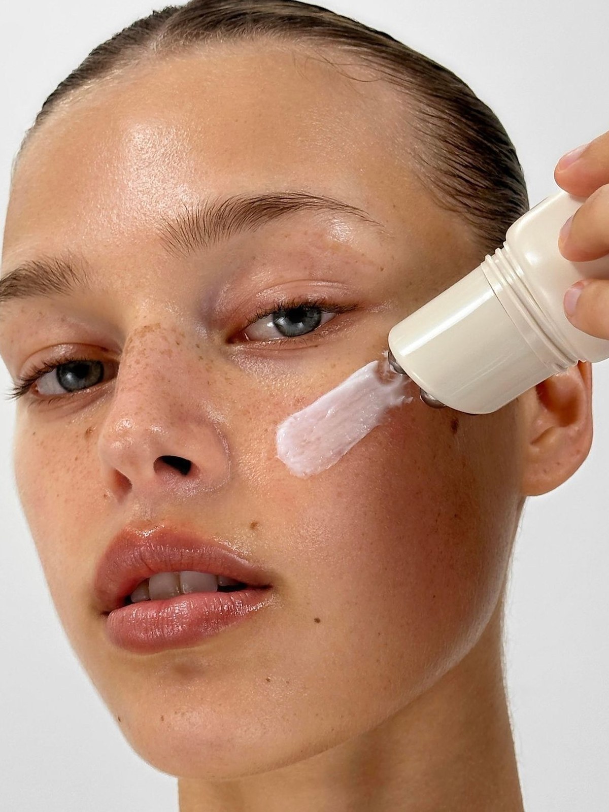Woman with slicked-back hair and freckles applying a pale pink cream to her cheek from a rounded white bottle
