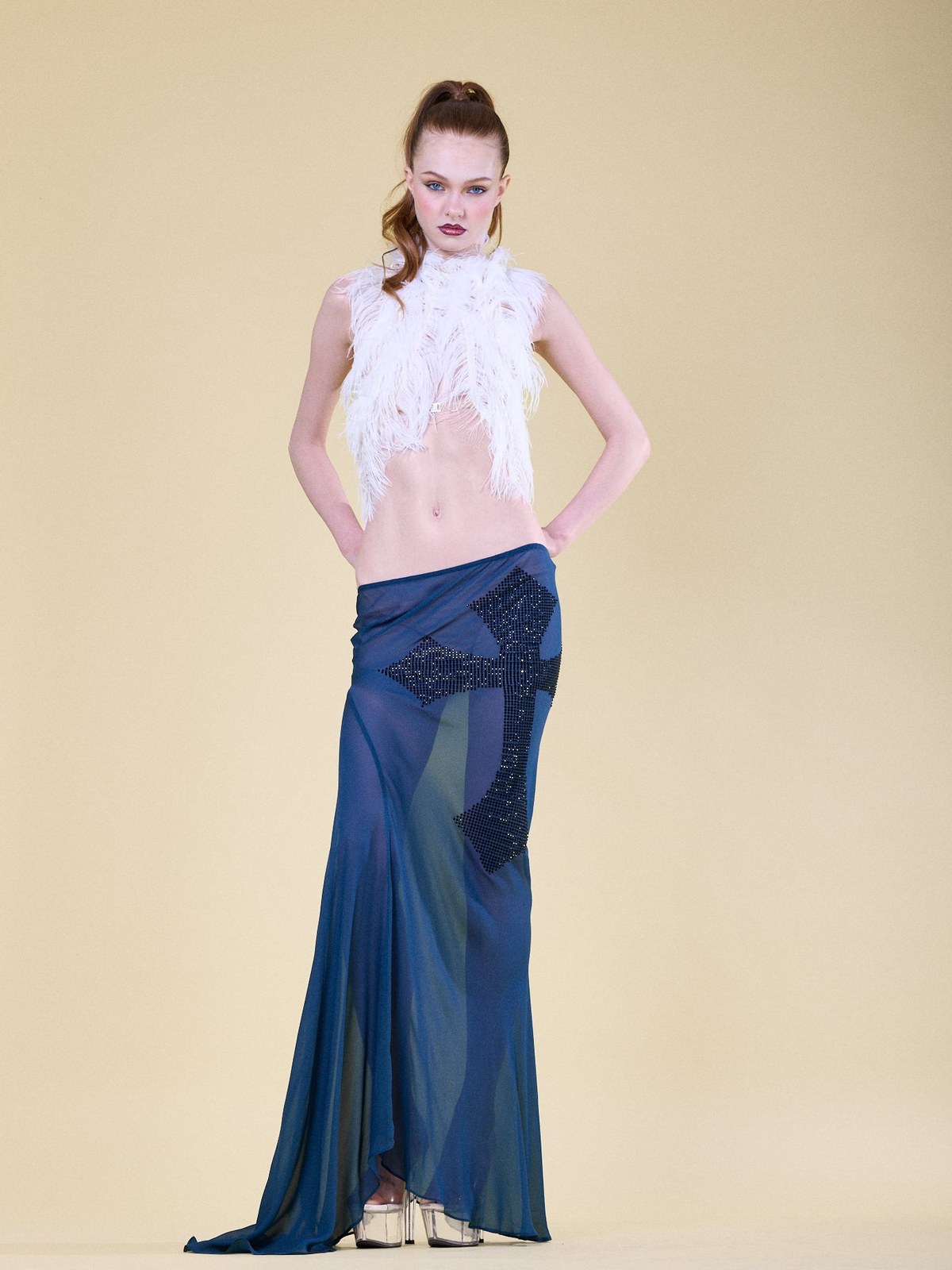 Model wearing a white feathered top and blue skirt against a warm neutral studio backdrop