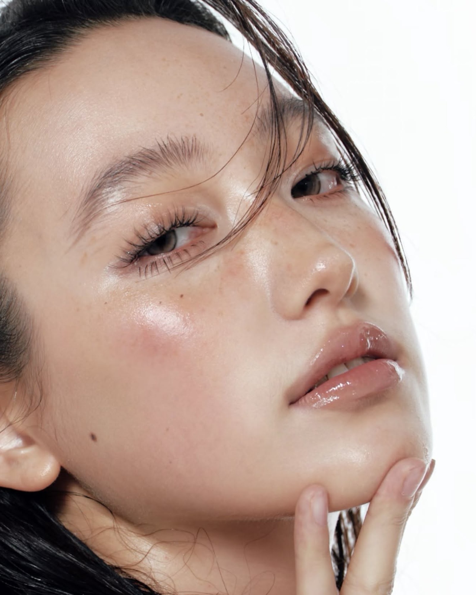 Close-up of a woman with wet dark hair, freckles and glossy lips tilting her chin on her fingertips