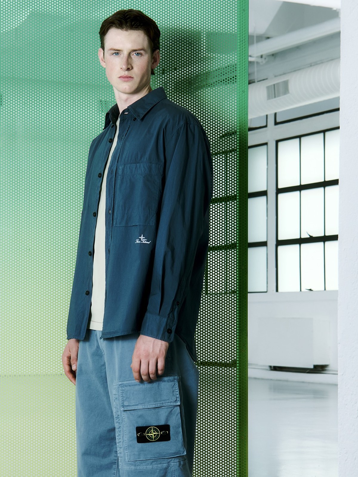 Model in a navy overshirt and light blue cargo trousers against a green gradient backdrop