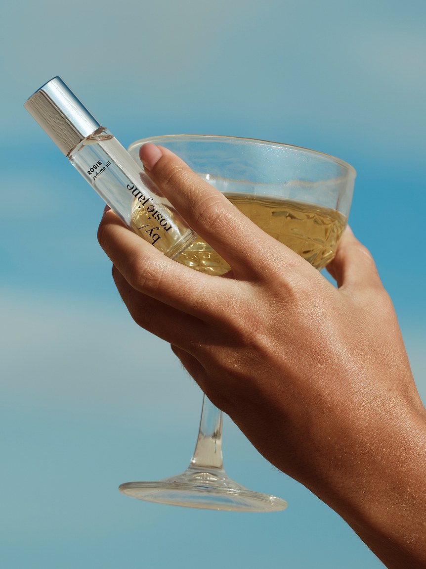 A hand holding By Rosie Jane fragrance beside a drink against a blue backdrop