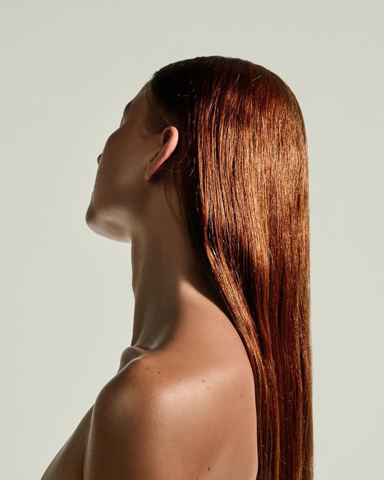 Side profile of a woman with long, straight, glossy copper red hair and bare shoulders against a pale grey backdrop