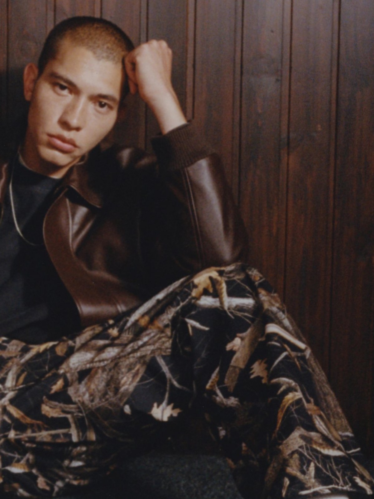 Male model seated in a brown leather jacket and camo pants against wood paneling