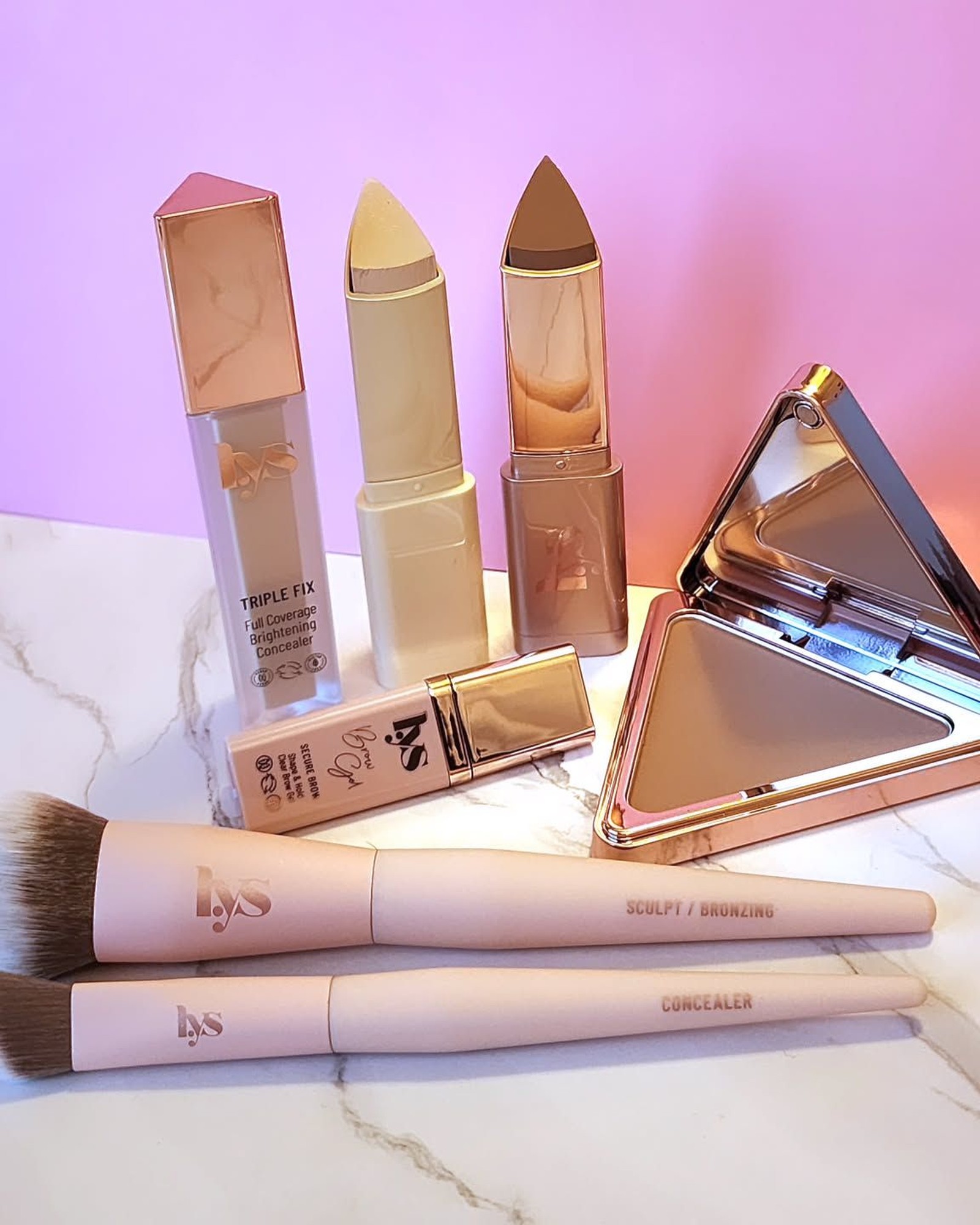 LYS Beauty makeup arranged on white marble against a pink backdrop: a concealer tube, two cream sticks, a triangular bronzer compact and two pink brushes.