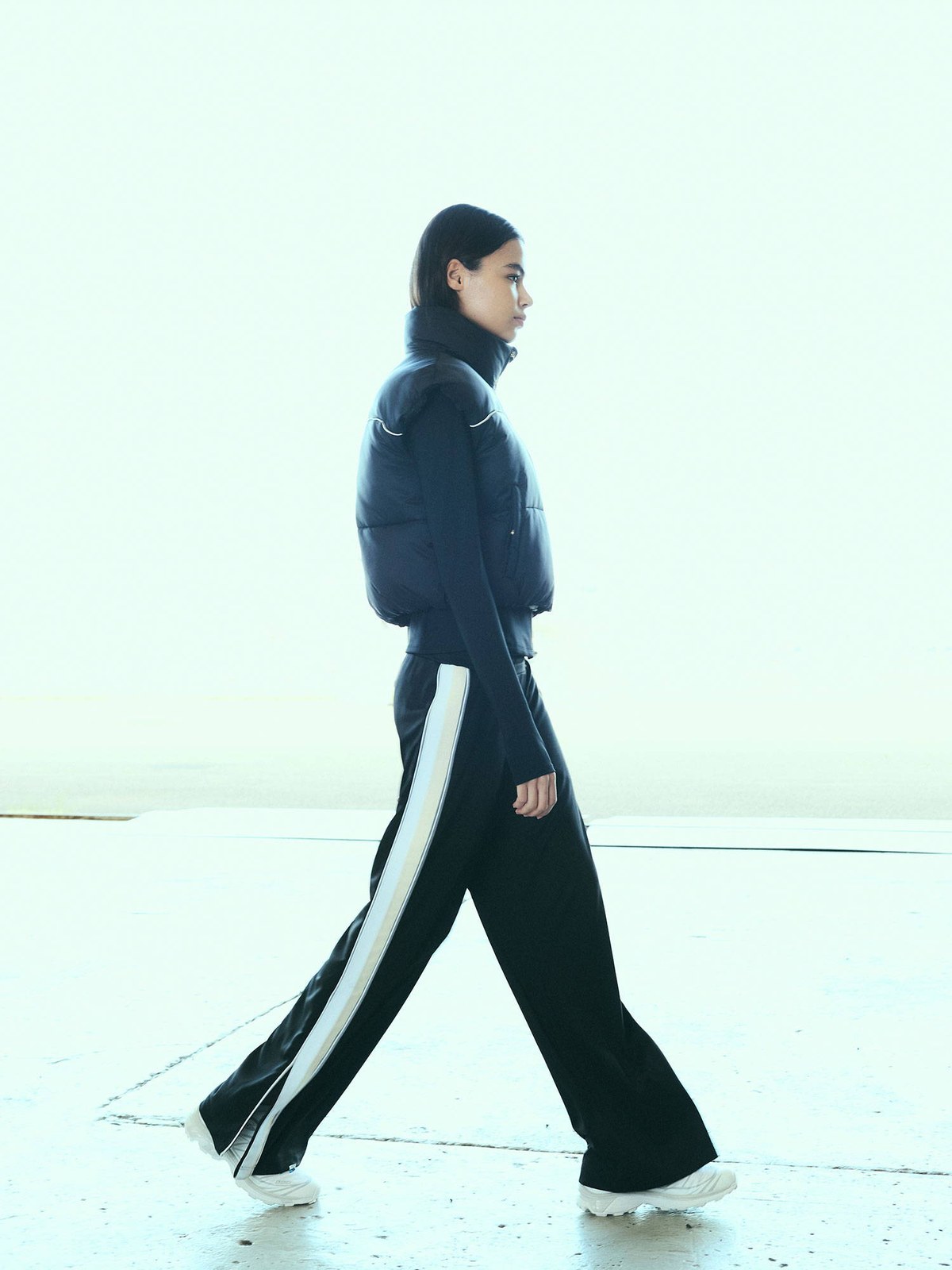 Model walking outdoors in a dark tracksuit beside an aircraft