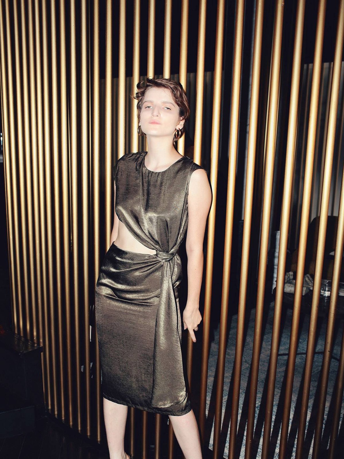 Model in a sculptural silver-gray dress against golden vertical slats