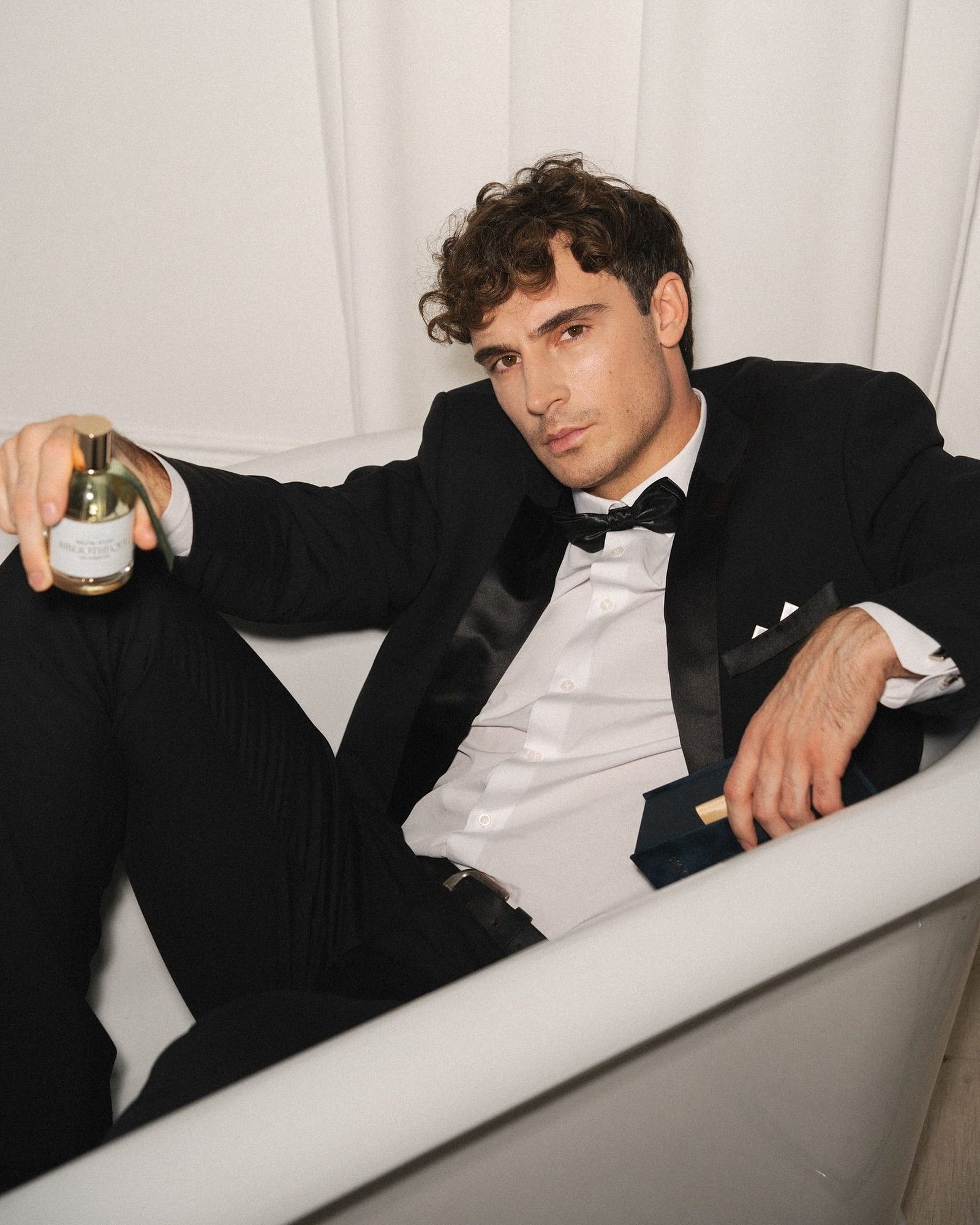 Man with curly brown hair in a black tuxedo and bow tie lounging in a white bathtub, holding a gold-capped fragrance bottle