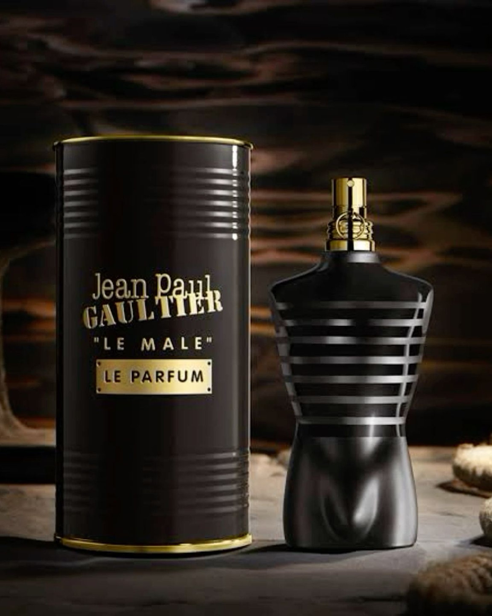 Jean Paul Gaultier Le Male Le Parfum black torso bottle with gold cap beside its black and gold tin, on a dark moody background